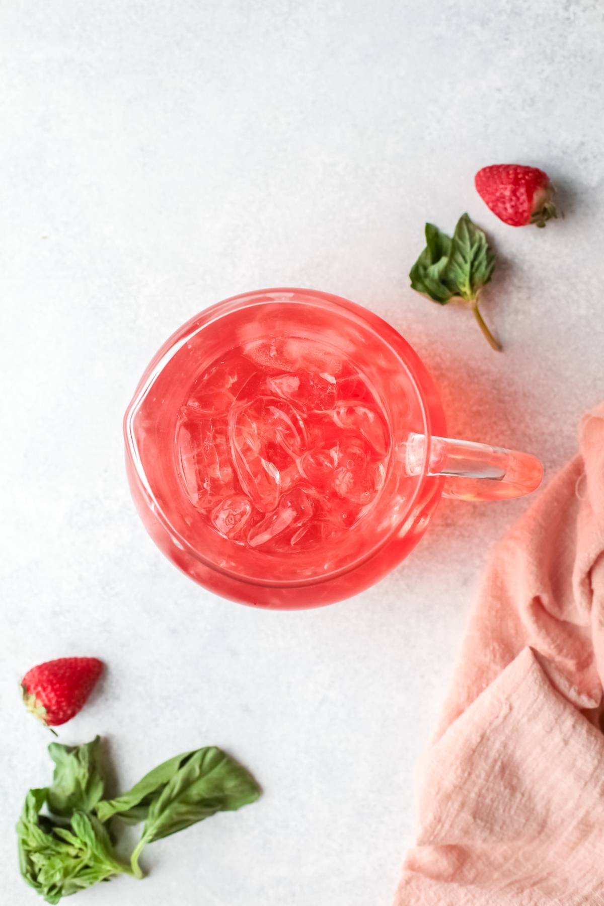 Pitcher of pink iced drink on a white surface, with strawberries, mint leaves, and a pink cloth nearby—a refreshing cocktail with gin and strawberry basil simple syrup, offering a vibrant variation on a classic gin rickey.