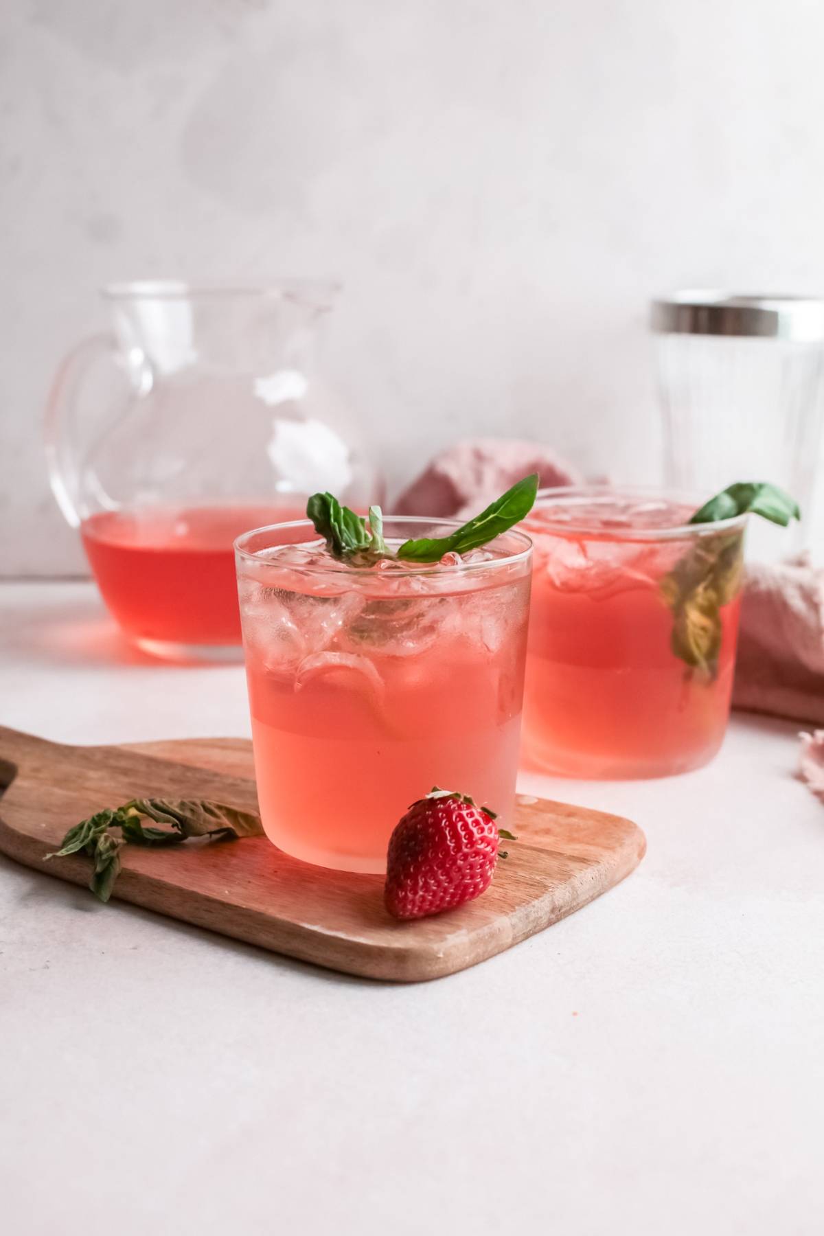 Two glasses of pink Strawberry gin rickey with ice, garnished with basil and a fresh strawberry, sit on a wooden board—a refreshing variation on a classic gin rickey featuring strawberry basil simple syrup.