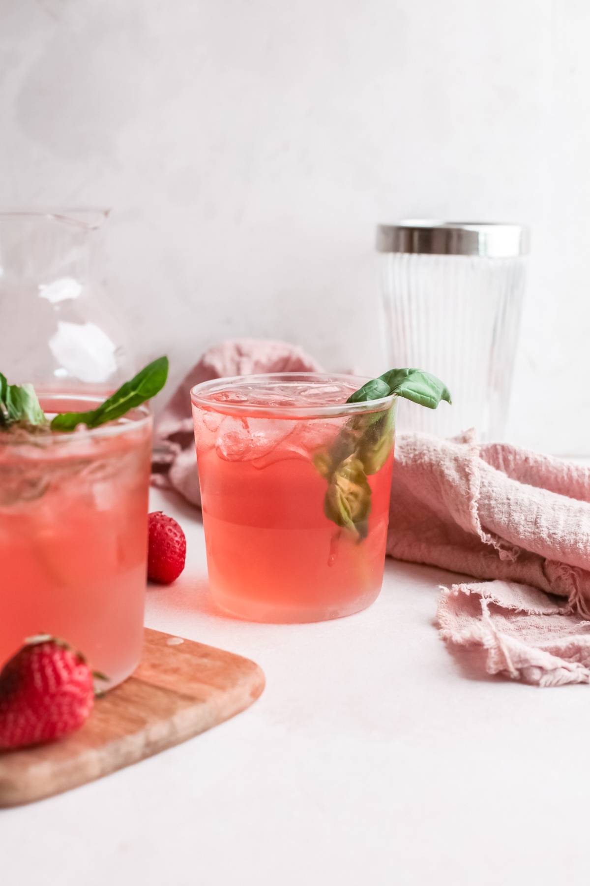 A glass of pink strawberry basil drink with ice, garnished with basil, on a light surface with fresh strawberries—a refreshing variation on a classic gin rickey made with strawberry basil simple syrup.