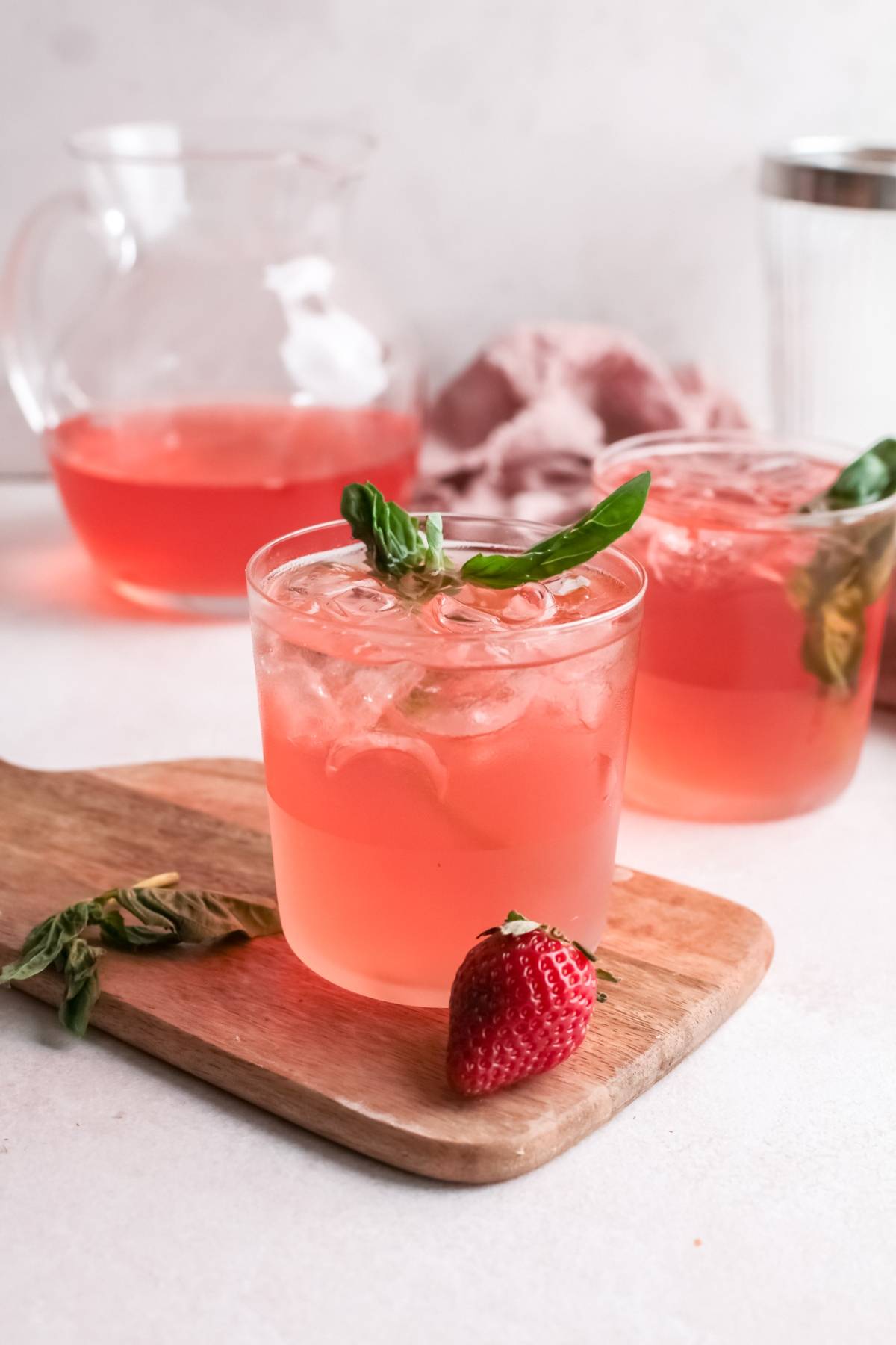 Two glasses of pink iced drink with basil, a fresh strawberry, and a pitcher in the background on a wooden board—perfect for serving as a refreshing summer cocktail or Strawberry gin rickey made with strawberry basil simple syrup.
