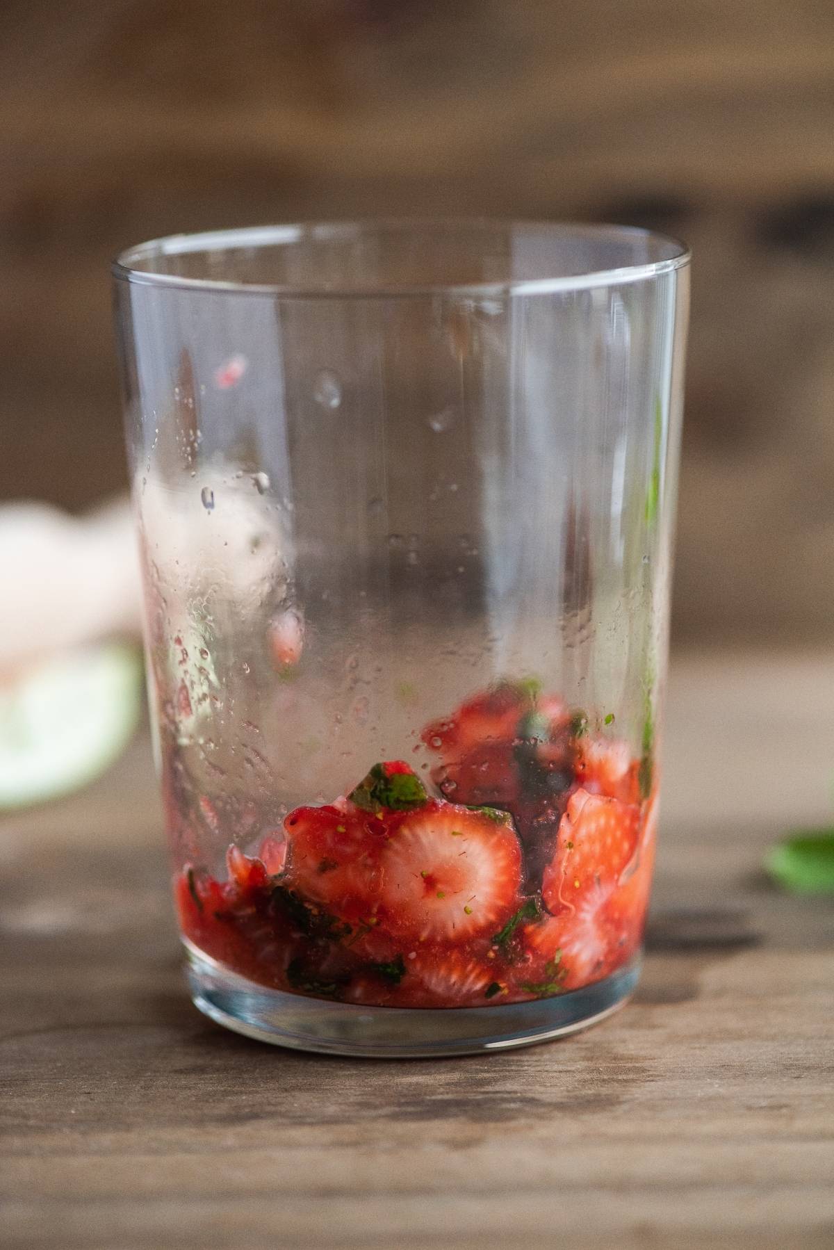 Glass with muddled strawberries and mint on a wooden surface, partially filled, capturing a vibrant drink with strawberries, set against a blurred background.