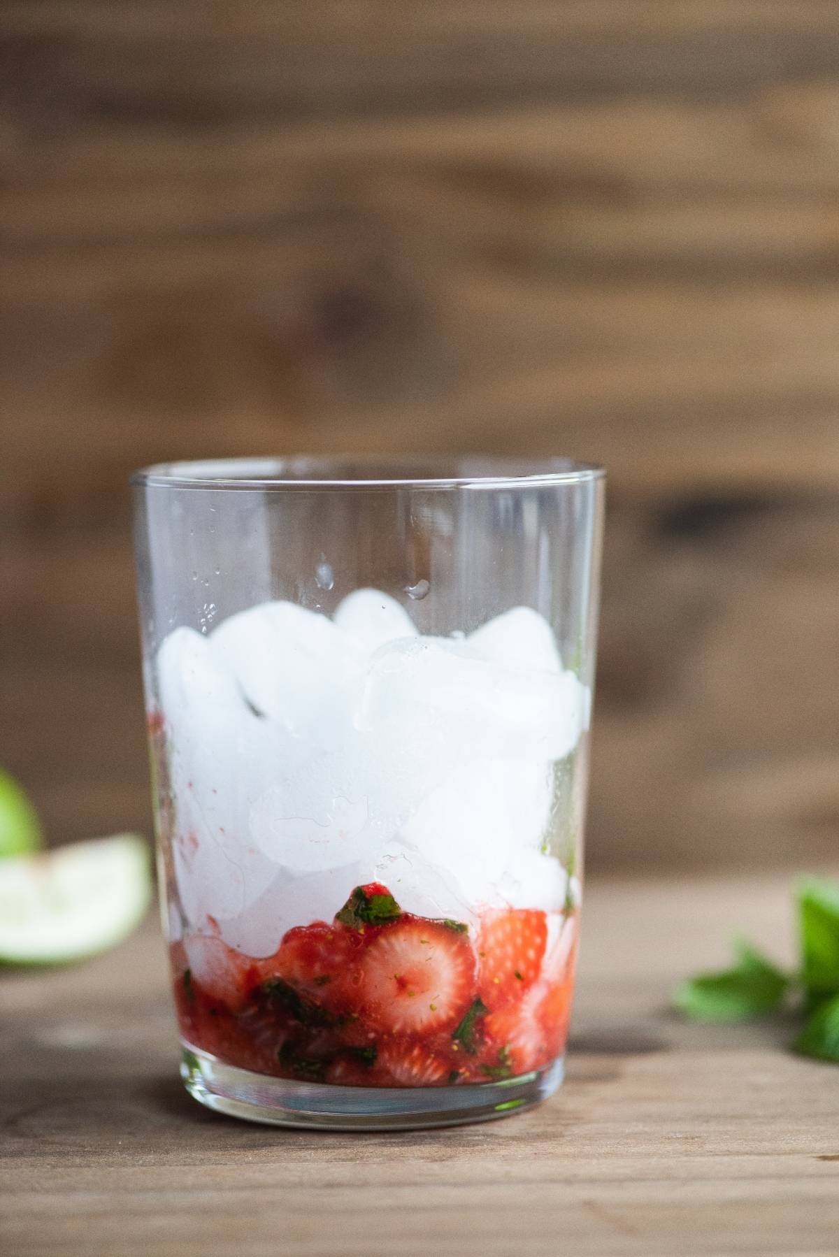 A glass with ice, chopped strawberries, and herbs sits on a wooden surface, ready to become a refreshing strawberry mojito cocktail with white rum or even Cachaça for a twist, with lime and greens nearby.