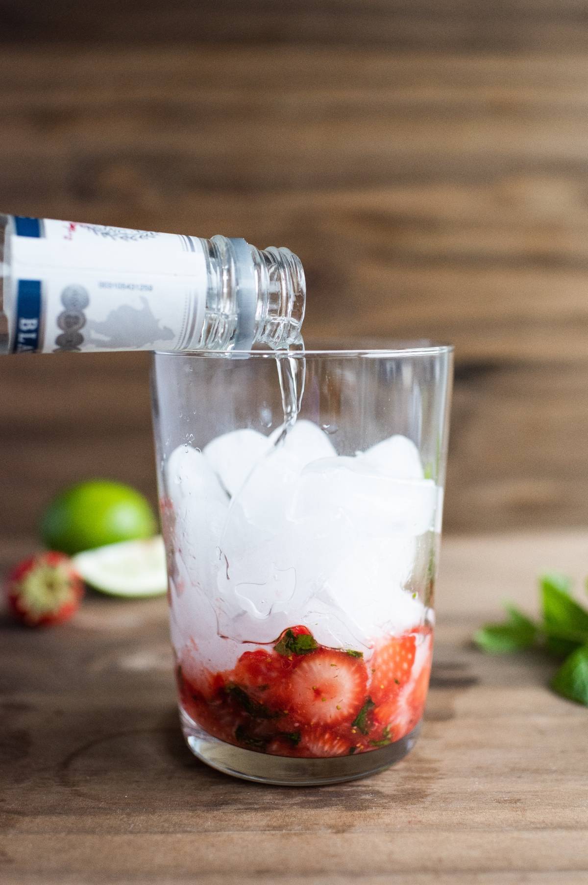 Clear liquor being poured into a glass with ice, sliced strawberries, and mint; lime in background—an easy summer cocktail recipe for a refreshing strawberry mojito.