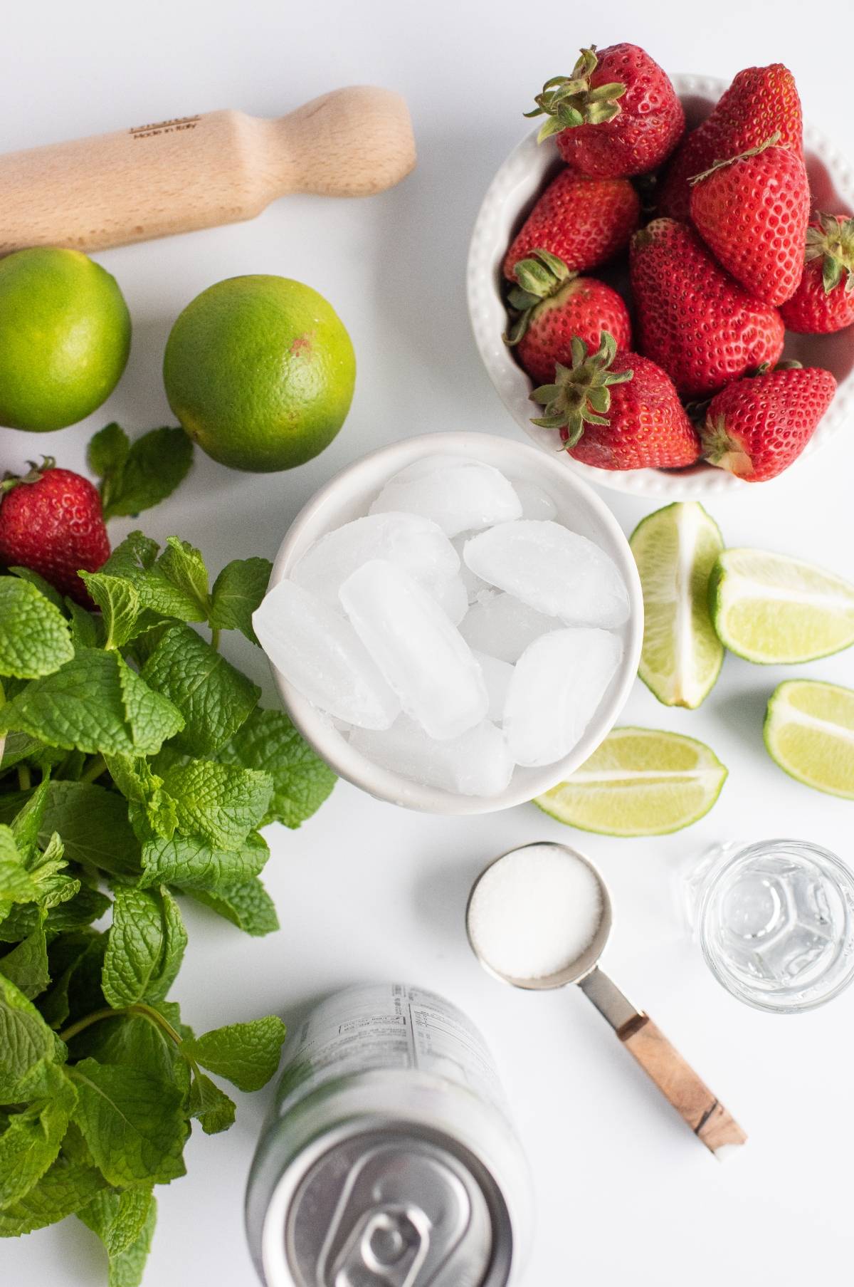 Fresh strawberries, limes, mint, ice, sugar, a jigger of clear liquid—everything you need for an easy summer cocktail recipe. Create a refreshing Strawberry mojito cocktail with white rum using these simple ingredients on a white surface.