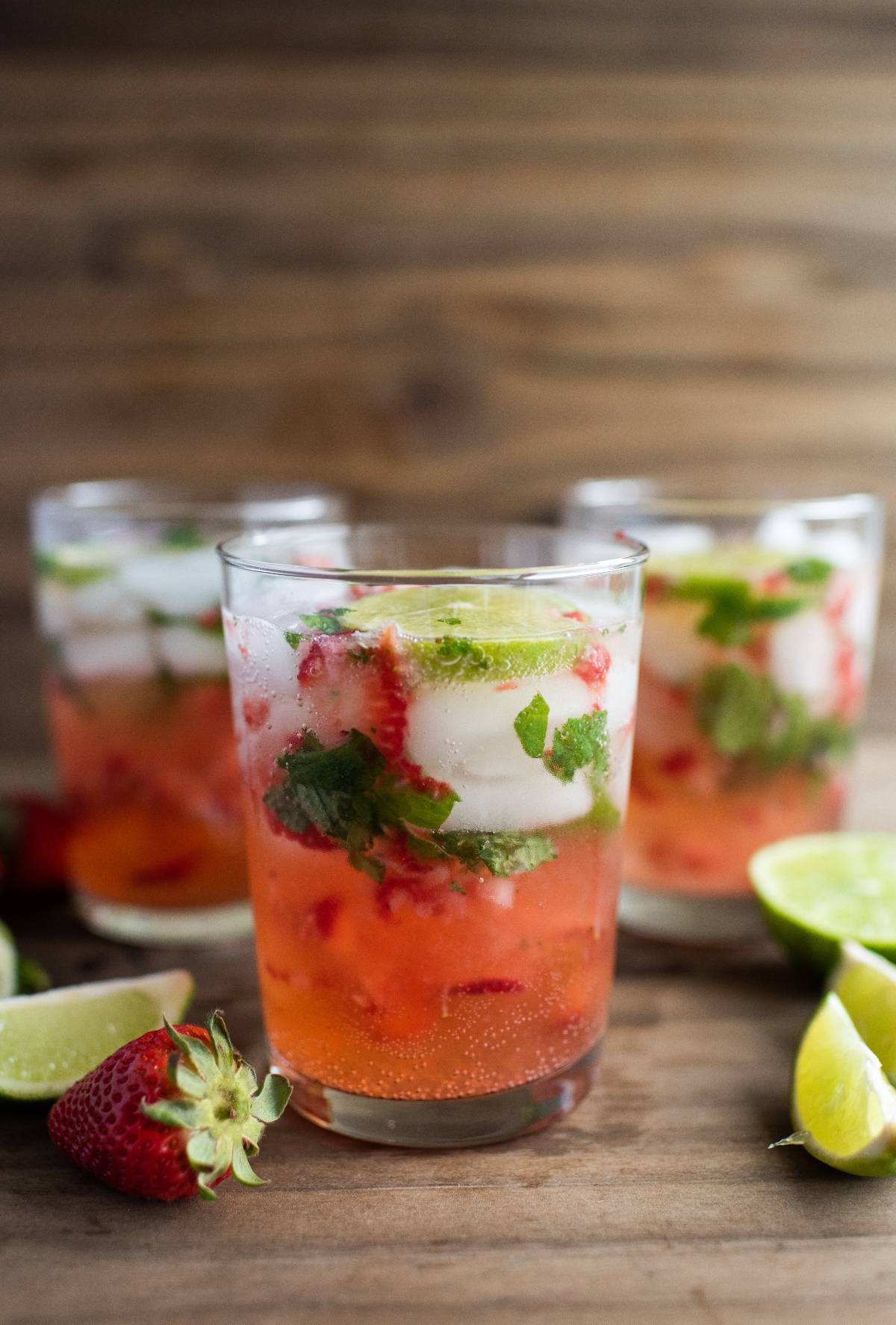 Three glasses of strawberry mojito, a refreshing cocktail with white rum, ice, mint, lime slices, and fresh strawberries sit on a wooden surface—an easy summer cocktail recipe perfect for warm days.