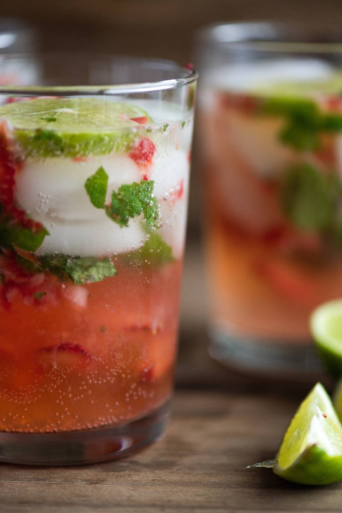 A glass of strawberry and mint cocktail with white rum, ice, and lime slices, with another glass and lime wedges in the background—an easy summer cocktail recipe to try.