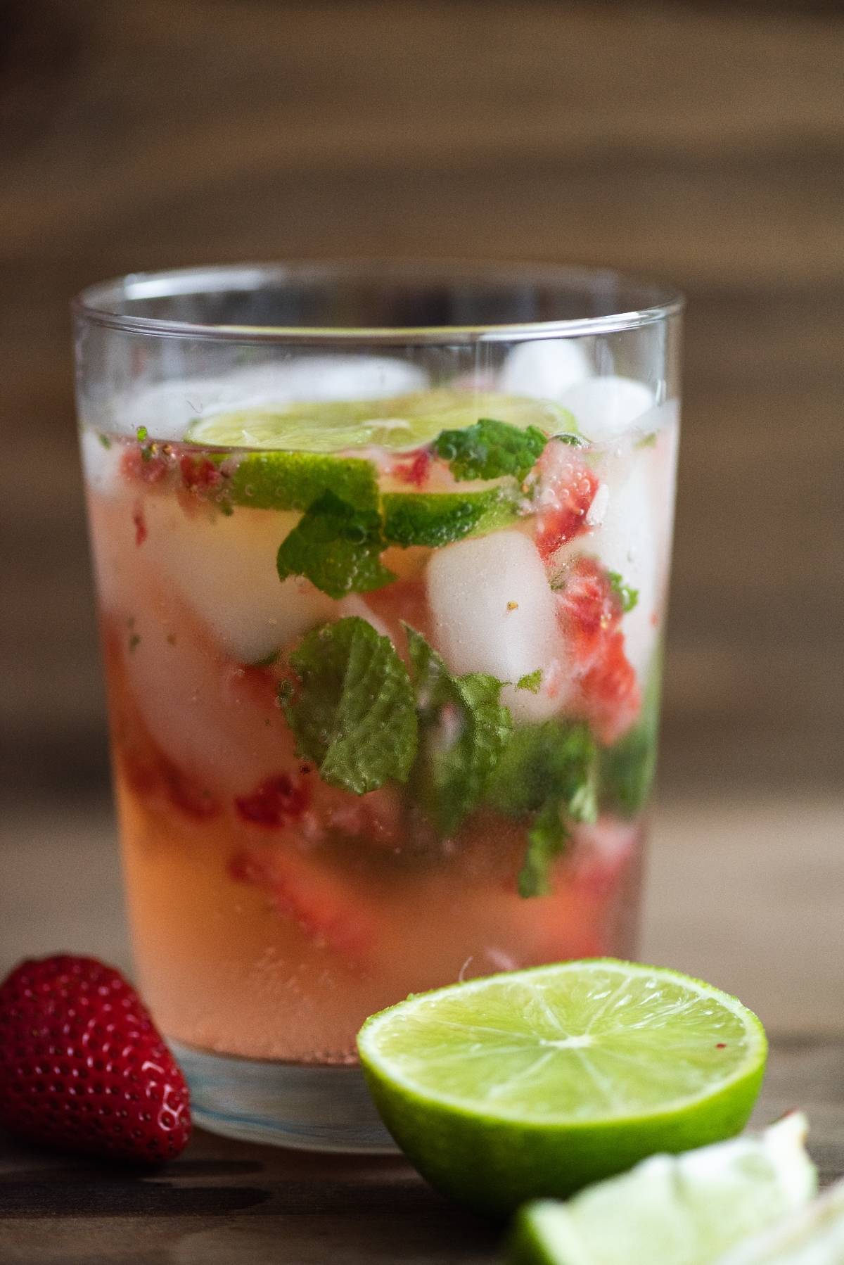 A glass of strawberry mojito cocktail with white rum, served with ice, mint leaves, and lime, garnished with fresh strawberries and lime in front.