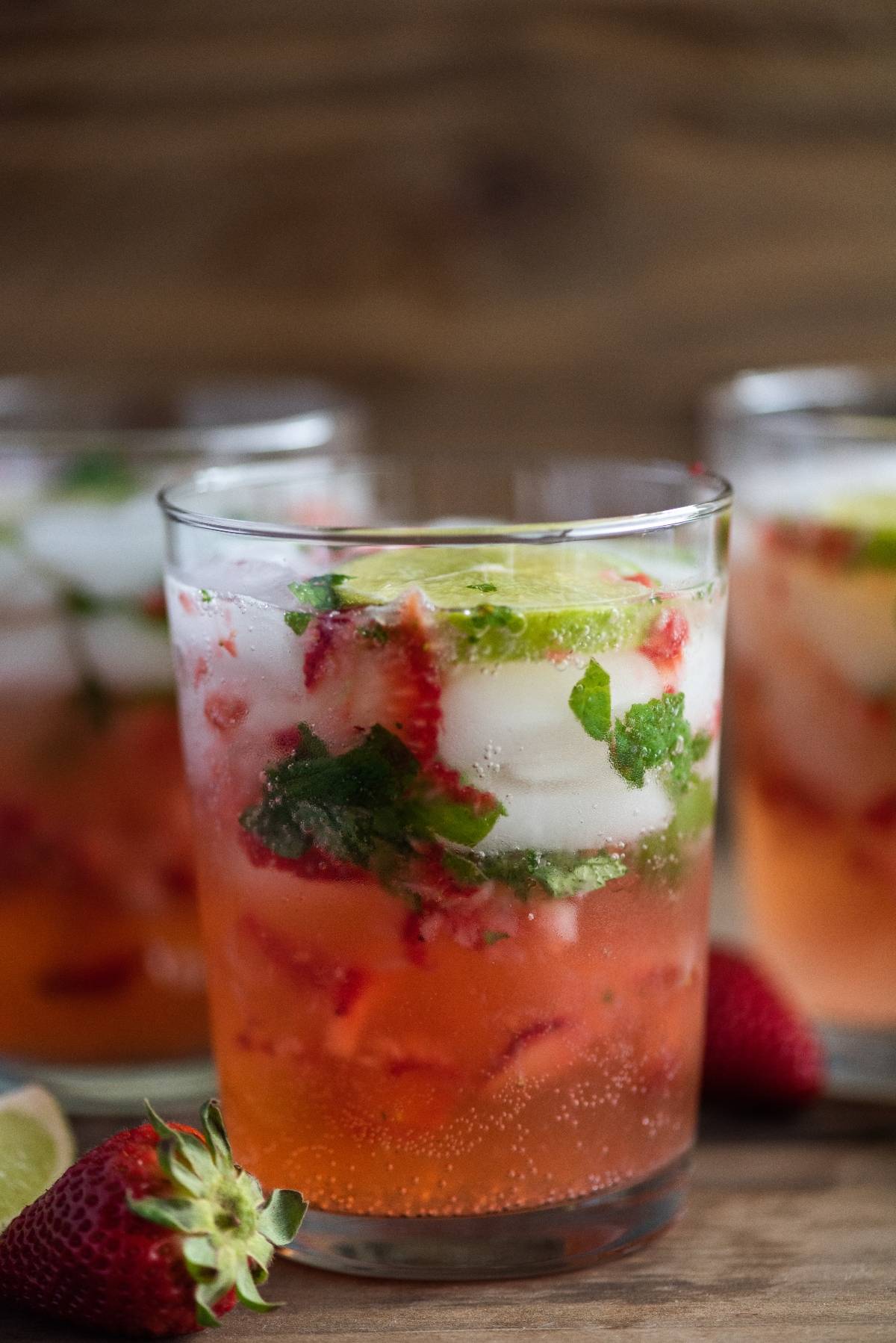 A refreshing strawberry mojito—an easy summer cocktail recipe—features ice, mint, lime slices, and fresh strawberries, with two more glasses behind.