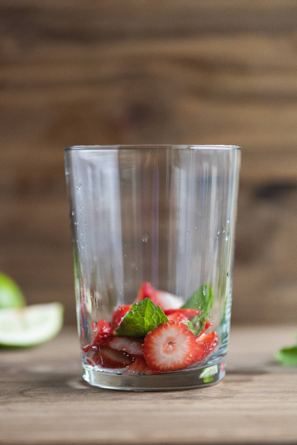 A clear glass of Strawberry Mojito, garnished with sliced strawberries and mint leaves, sits on a wooden surface with a blurred background—an easy summer cocktail recipe perfect for any occasion.