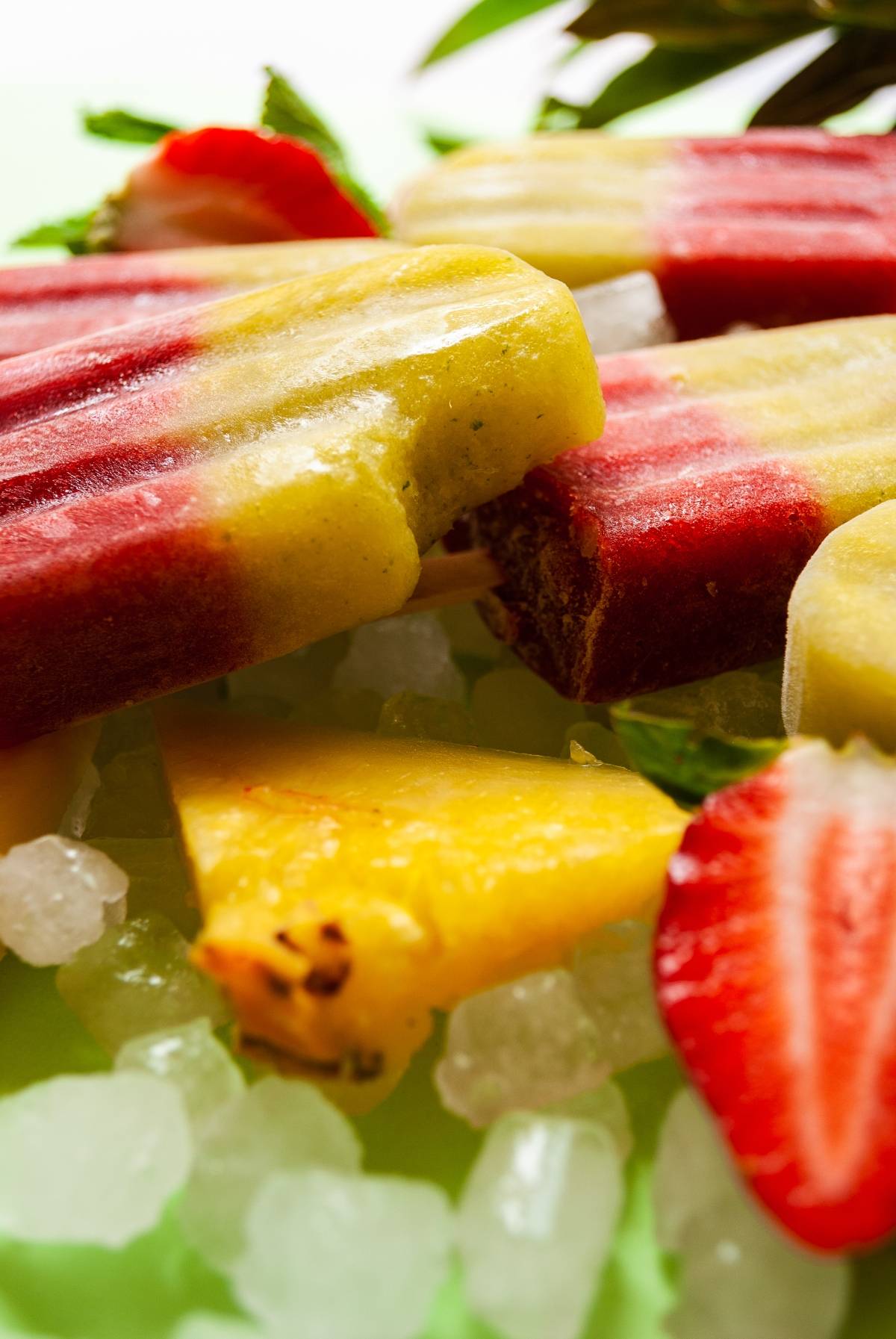 Colorful fresh fruit popsicles with red and yellow layers, served on ice with fresh strawberry and pineapple slices—these refreshing ice pops are perfect for a summer treat.
