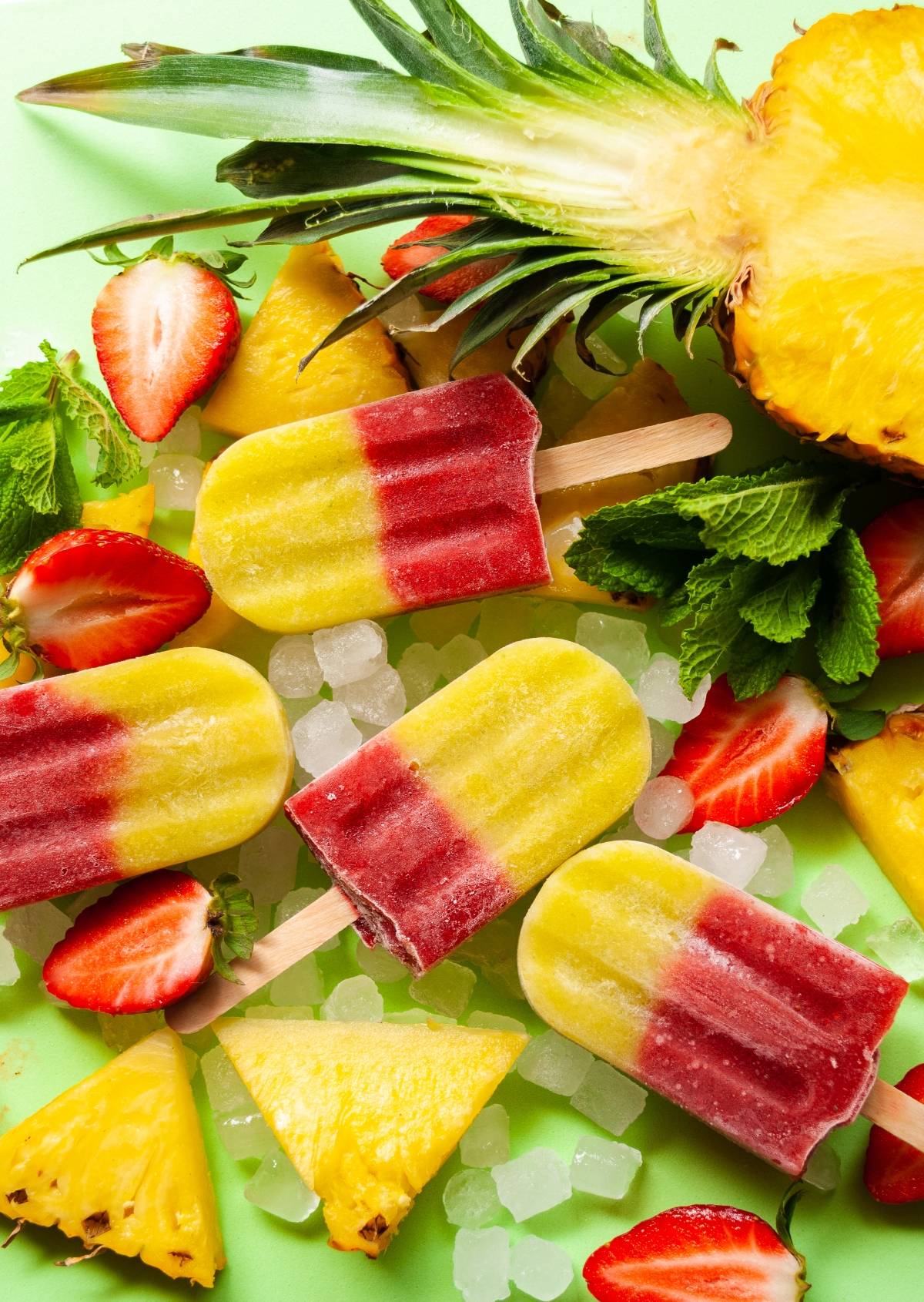 Colorful Strawberry Pineapple Popsicles on ice, surrounded by fresh pineapple slices, strawberries, and mint leaves—perfectly refreshing ice pops made with fresh fruit.
