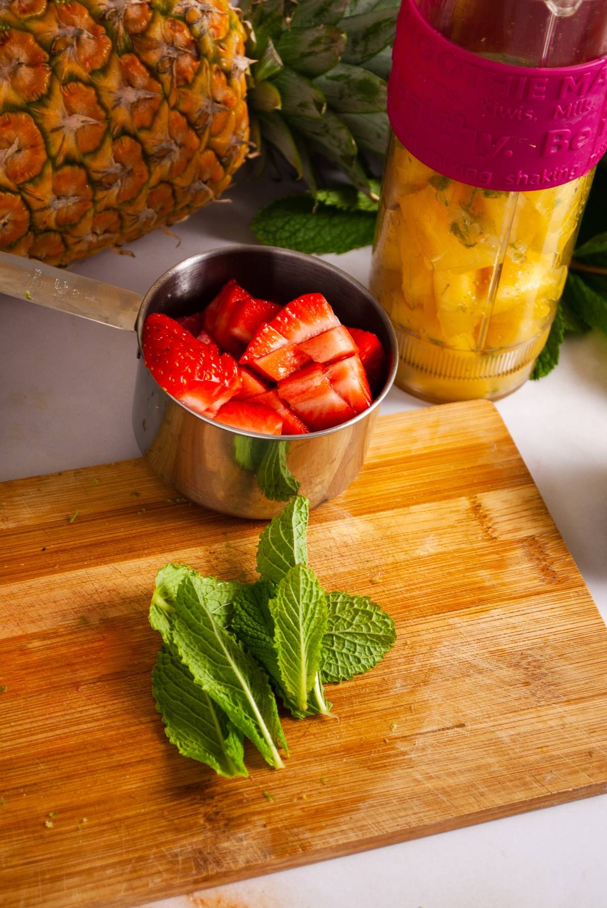 Chopped strawberries in a measuring cup, fresh mint leaves, pineapple, and a drink shaker on a wooden board—perfect ingredients for an easy layered popsicle recipe or making refreshing ice pops at home.
