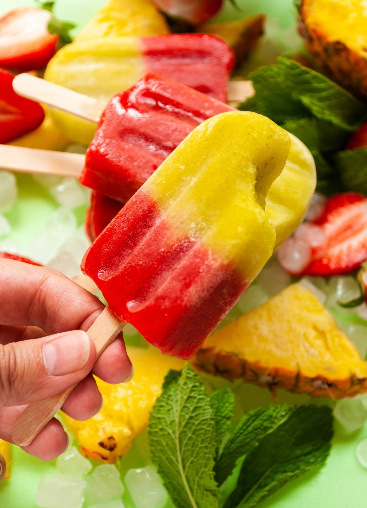 A hand holds a vibrant pineapple and mint popsicle over ice, fruit slices, and fresh mint leaves—an easy layered popsicle recipe for refreshing summer treats.