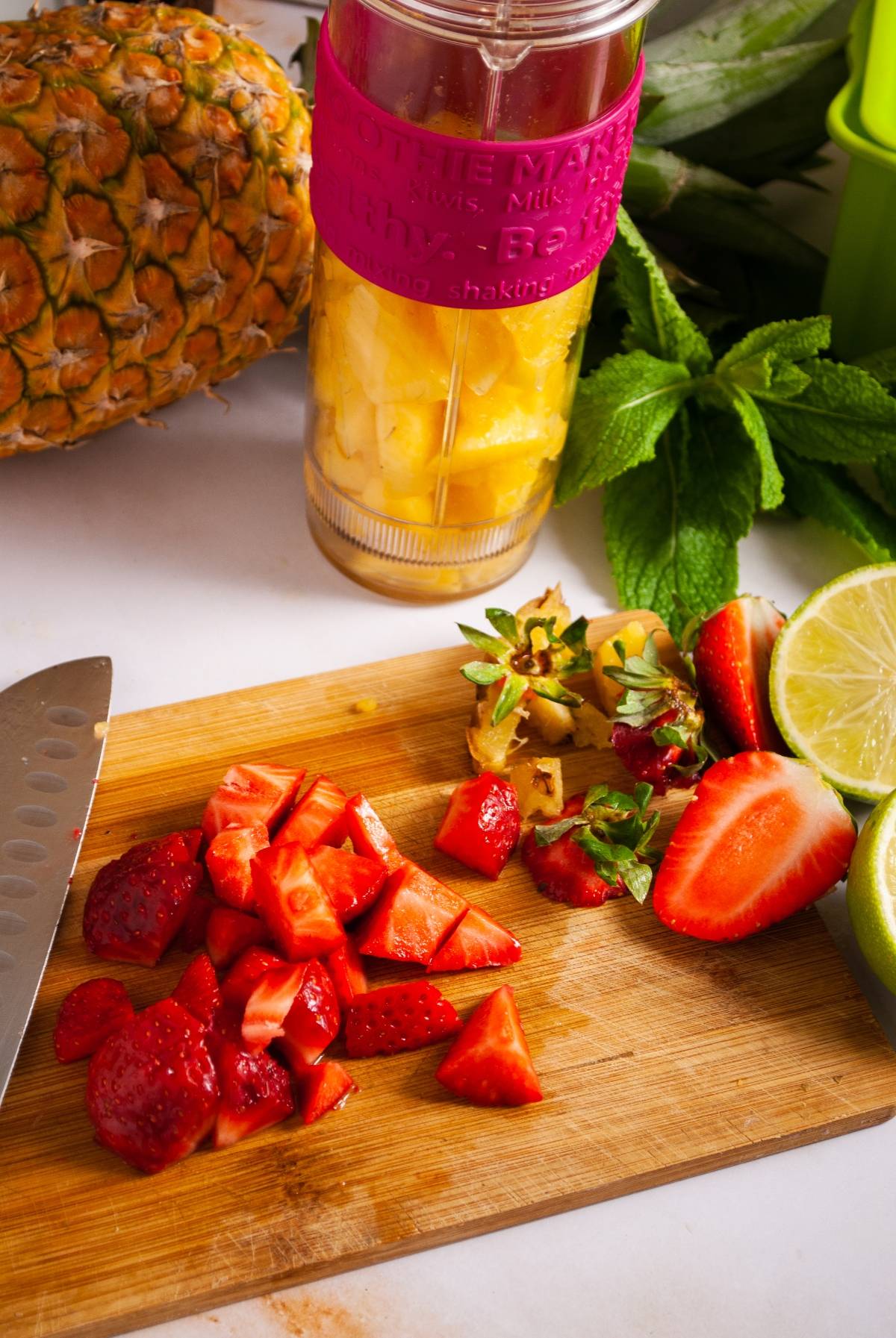 Chopped strawberries and lime on a cutting board, with pineapple and a blender in the background—perfect for making Strawberry Pineapple Popsicles or other refreshing ice pops.