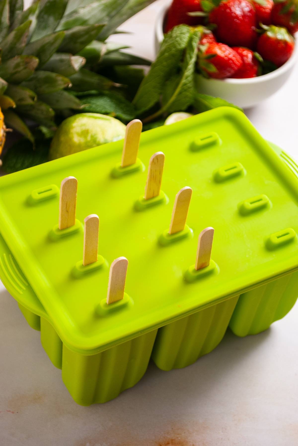 Green popsicle mold with wooden sticks, surrounded by fresh fruit and mint on a white surface—perfect for making a refreshing ice pops or Strawberry Pineapple Popsicles at home.