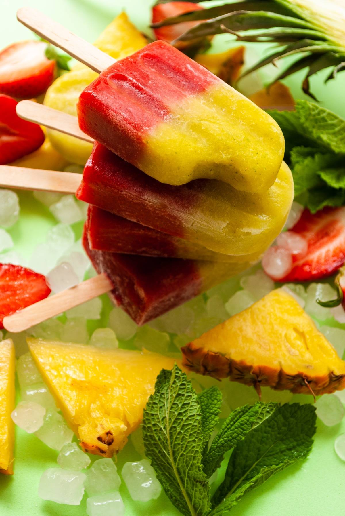 Colorful Strawberry Pineapple Popsicles stacked on ice, surrounded by strawberries, pineapple slices, and fresh mint leaves.
