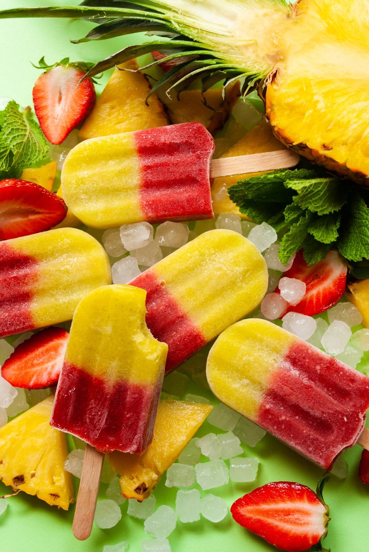 Colorful yellow and red fresh fruit popsicles on ice, surrounded by pineapple slices, strawberries, and fresh mint leaves for a refreshing treat.