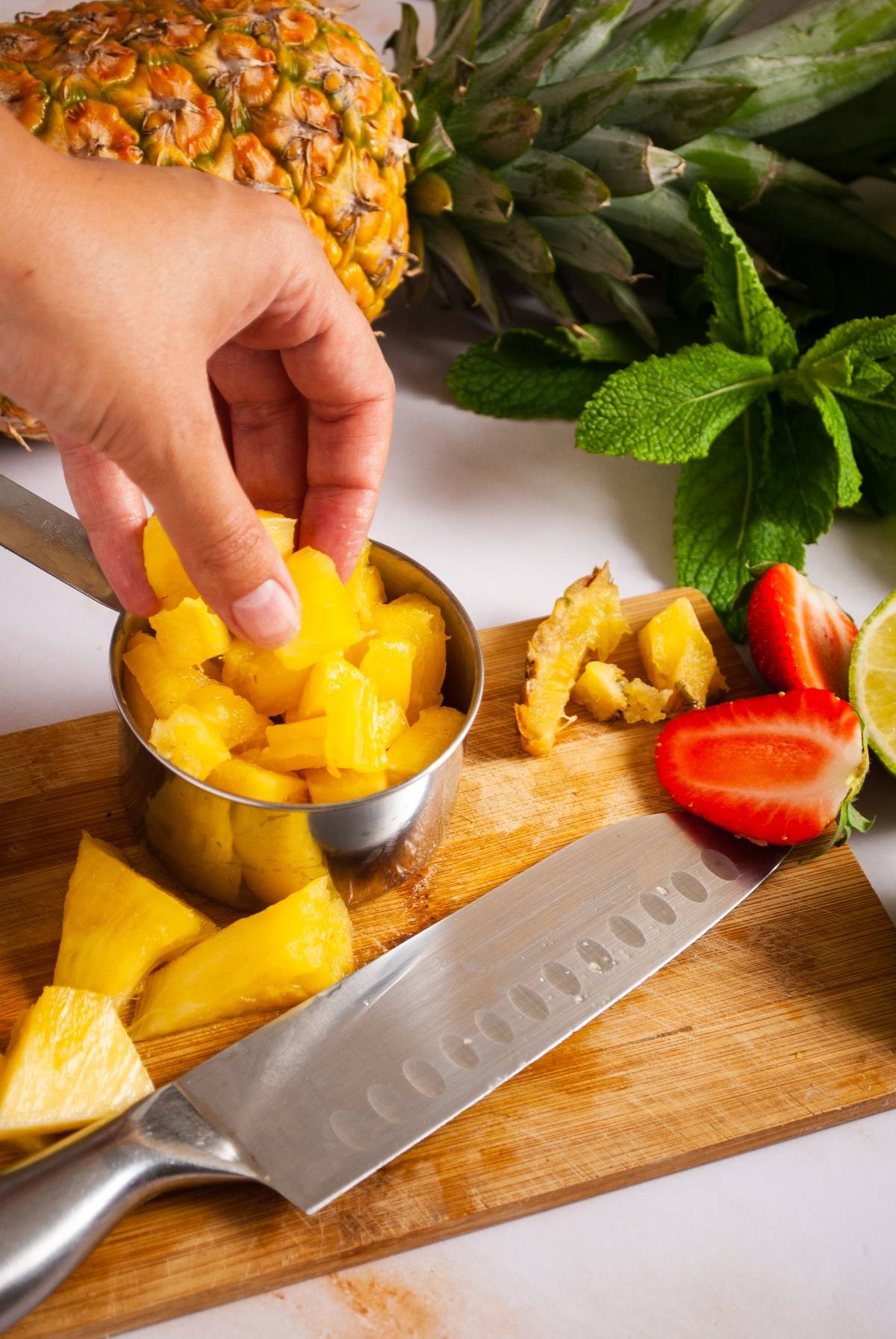 A hand places pineapple chunks into a metal cup on a cutting board with fruit, knife, and mint leaves nearby—perfect prep for an easy layered popsicle recipe.