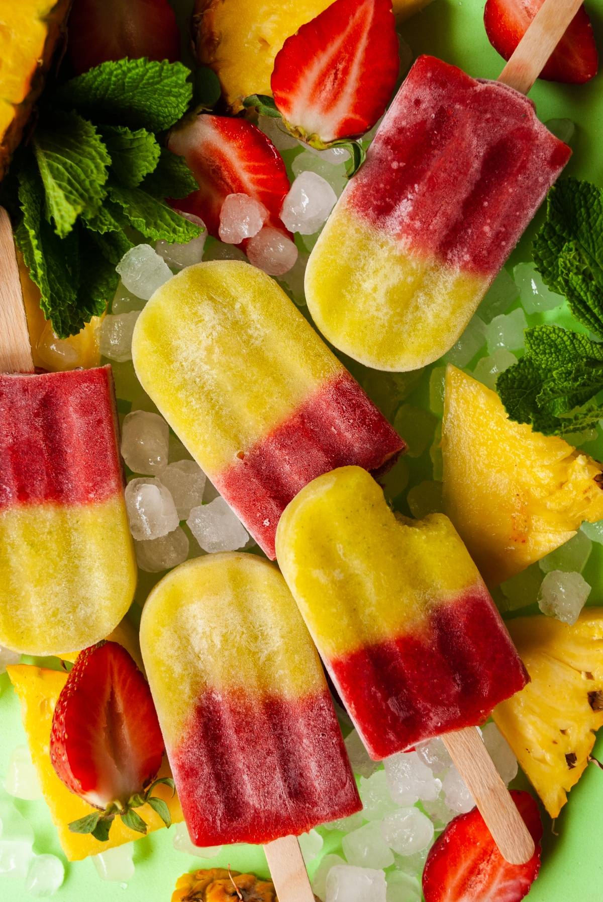 Colorful yellow and red popsicles made with strawberries, pineapple and mint, plus ice cubes on a green background—an easy layered popsicle recipe for refreshing ice pops.