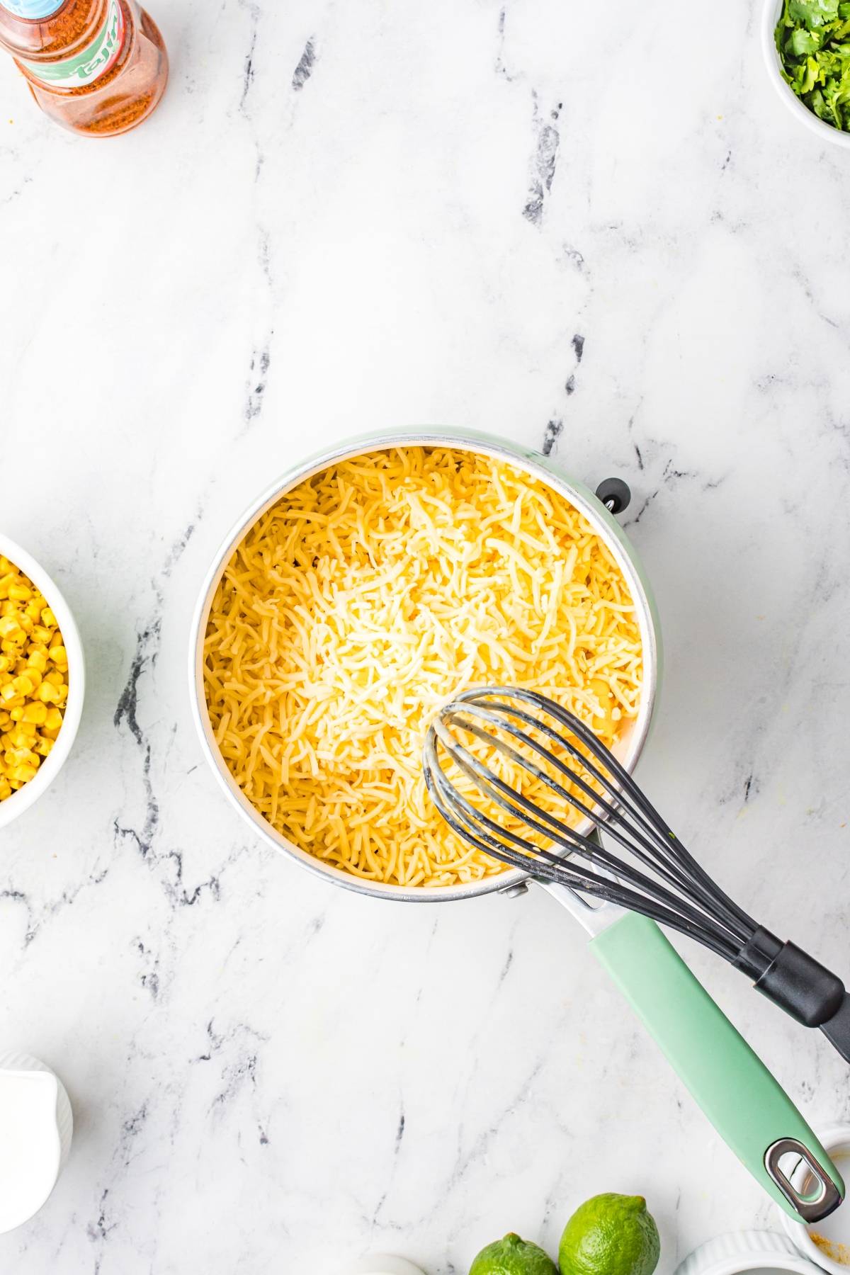 A pot of shredded cheese with a whisk on top, surrounded by bowls of corn, herbs, and limes—perfect ingredients for crafting Street Corn Nachos on a marble surface.