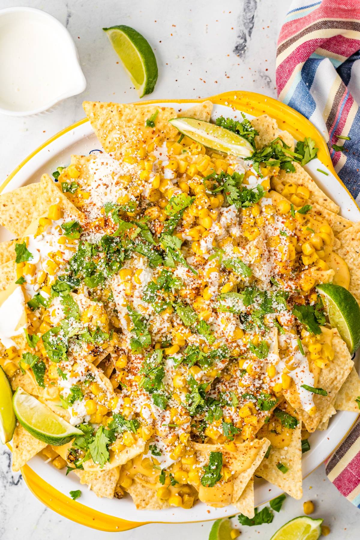 Tortilla chips topped with corn, cheese, cilantro, crema, and lime wedges bring Elote and nacho chips together for a Mexican nachos recipe with unique nacho toppings on a white plate.