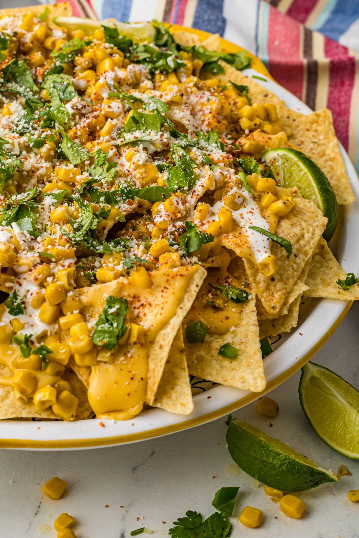 A plate of Street Corn Nachos topped with corn, cheese, cilantro, crema, and lime wedges on the side.