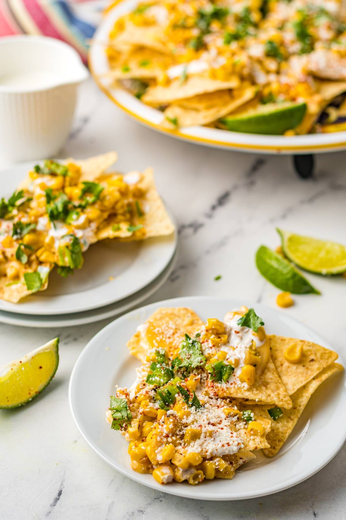 Tortilla chips topped with corn, cheese, sauce, and herbs create irresistible Street Corn Nachos—a Mexican nachos recipe with unique nacho toppings. Served on white plates with lime wedges nearby.