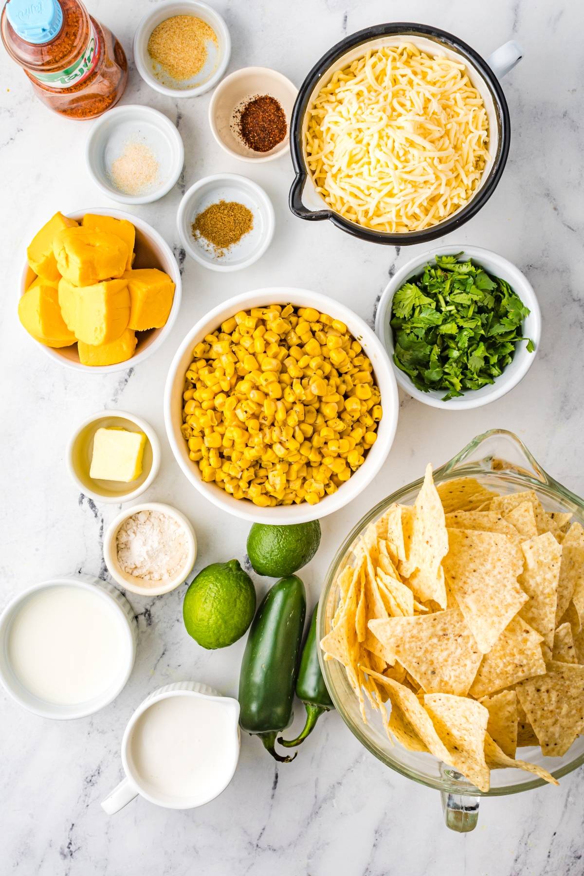 Bowls of corn, shredded cheese, chips, jalapeños, limes, cilantro, squash, butter, milk, and spices on a counter—perfect ingredients for delicious Street Corn Nachos or your favorite Mexican nachos recipe.