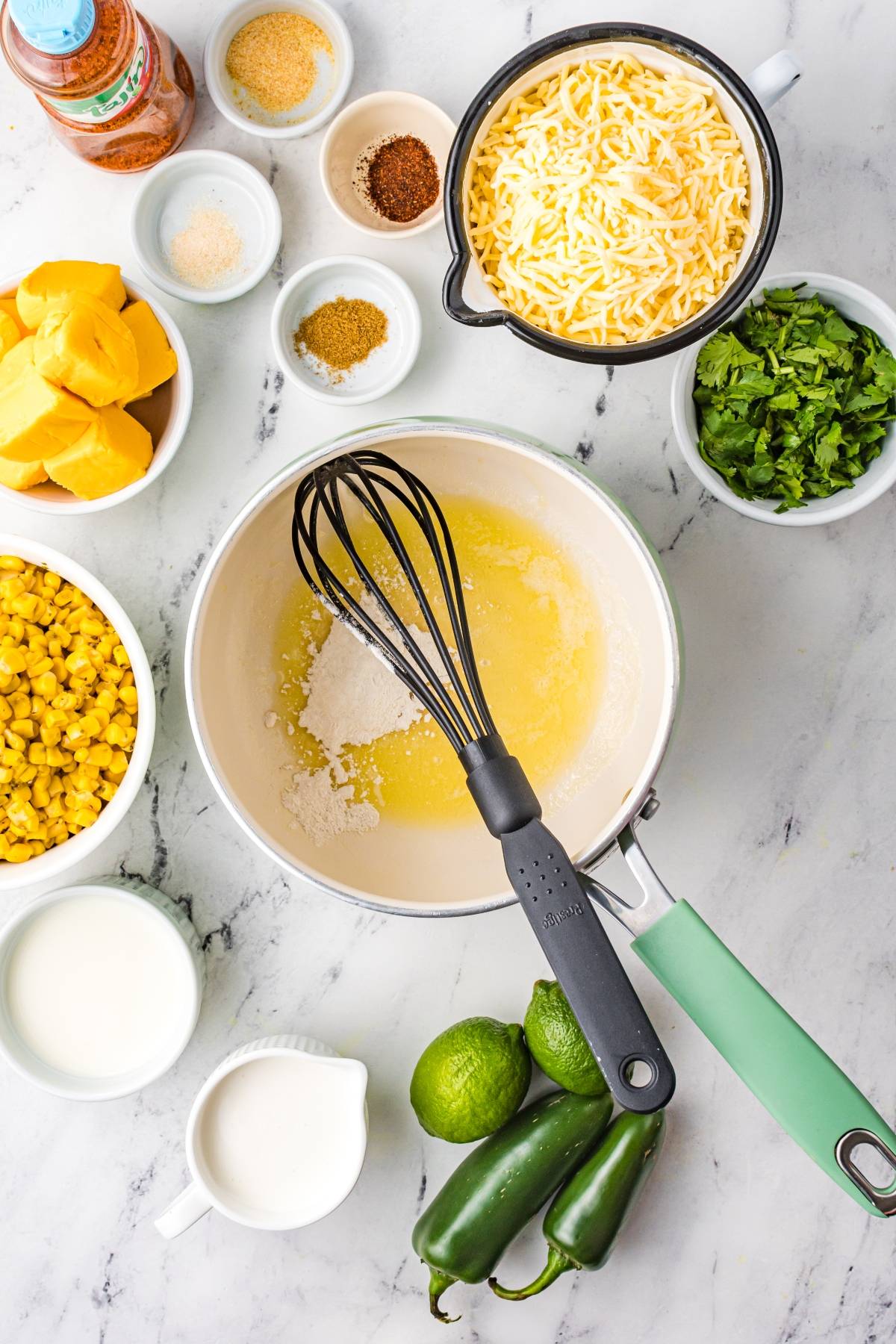A saucepan with melted butter and a whisk is surrounded by bowls of cheese, corn, seasonings, spinach, milk, and limes—perfect for creating unique nacho toppings or a delicious Nachos with Zesty Cheese Sauce.
