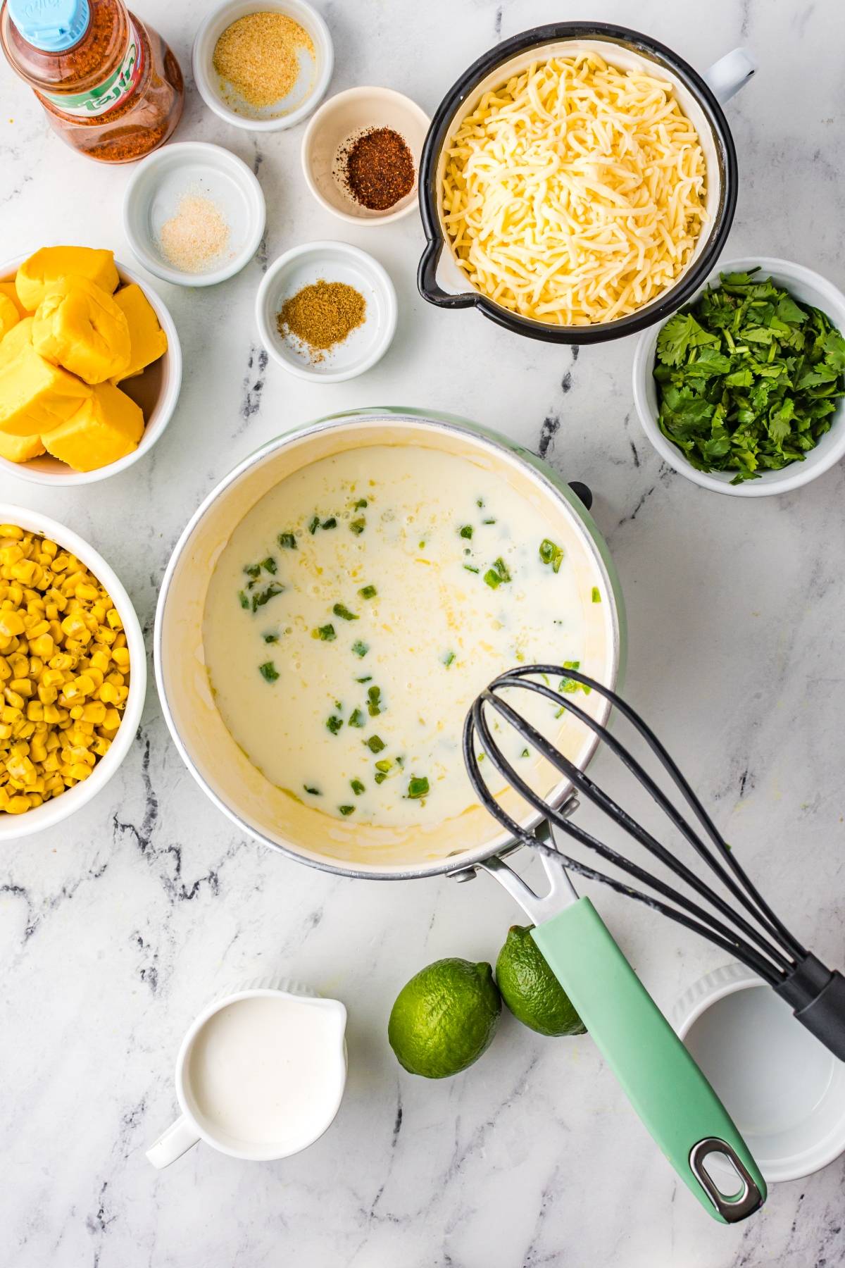 A saucepan of creamy sauce with chopped peppers, surrounded by cheese, spinach, corn, spices, and limes—perfect for street corn nachos or adding unique nacho toppings to your favorite Mexican nachos recipe.