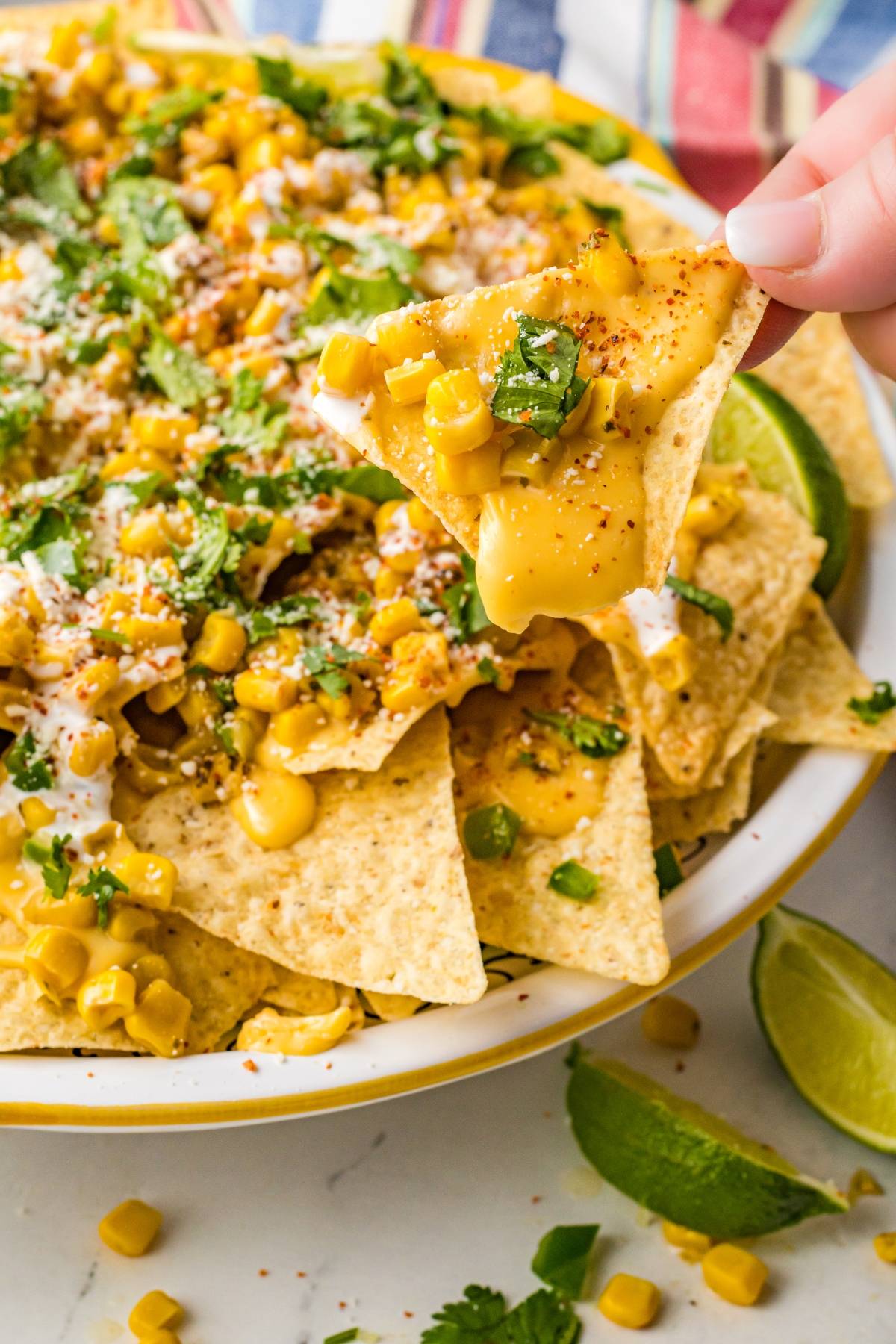 A hand holds a cheesy nacho chip topped with corn, cilantro, and seasoning above a plate of Street Corn Nachos with lime wedges—perfect for those craving unique nacho toppings.