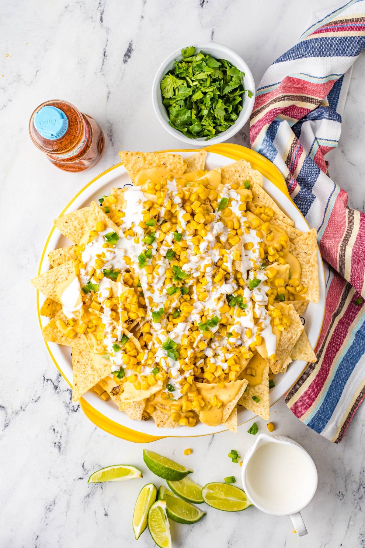 Plate of nachos with cheese, corn, sour cream, chopped greens, lime wedges, and cilantro on a marble table—an irresistible Mexican nachos recipe featuring unique nacho toppings for extra flavor.