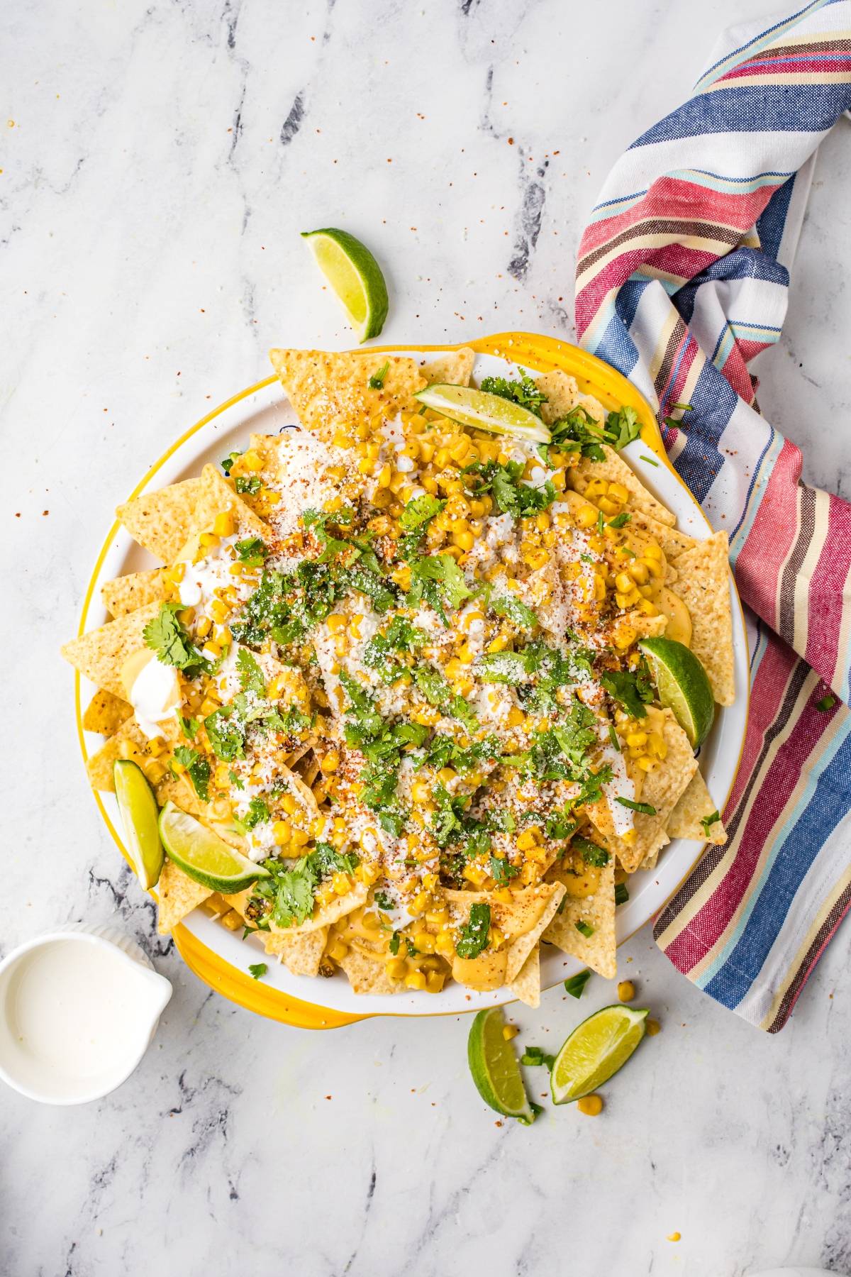 A plate of nachos topped with corn, cheese, cilantro, and lime wedges on a marble surface with a striped napkin—perfect for trying unique nacho toppings inspired by an Elote and nacho chips twist.