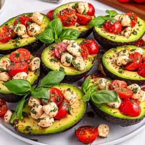 Caprese stuffed avocado halves topped with cherry tomatoes, mozzarella balls, fresh herbs, and basil leaves on a white plate.