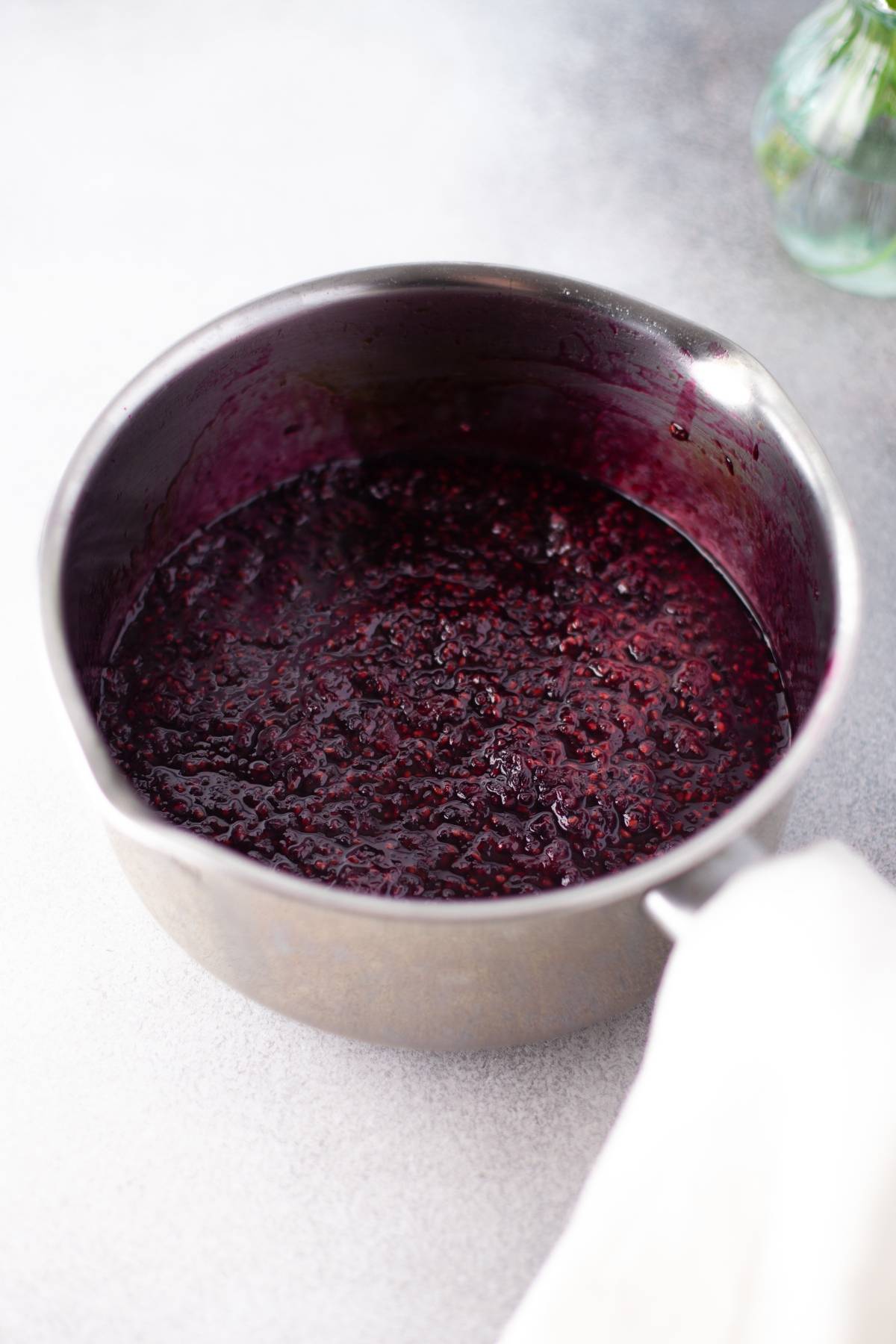 A saucepan filled with dark, simmered sugar free blackberry jam sits on a light countertop.