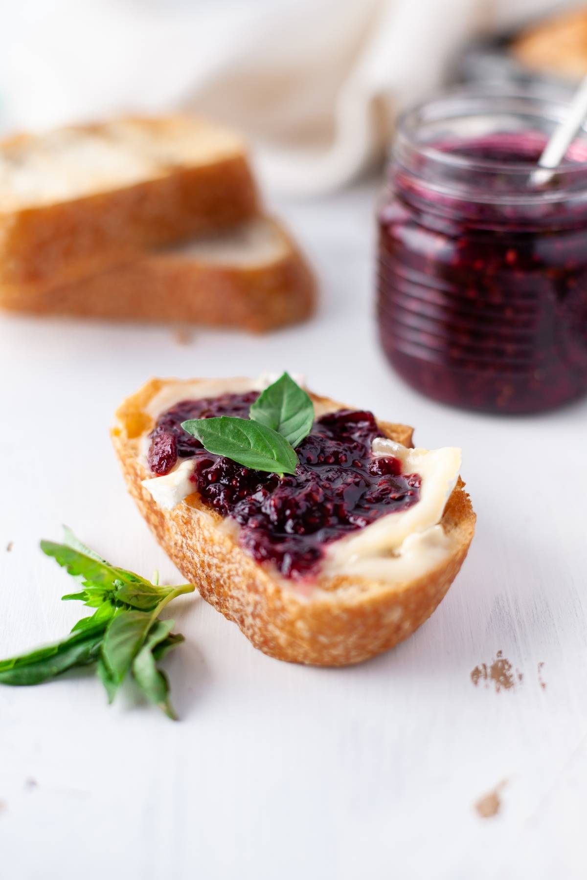 A slice of bread with butter and sugar free blackberry jam, garnished with a basil leaf, with a jar of jam made with fresh berries in the background.