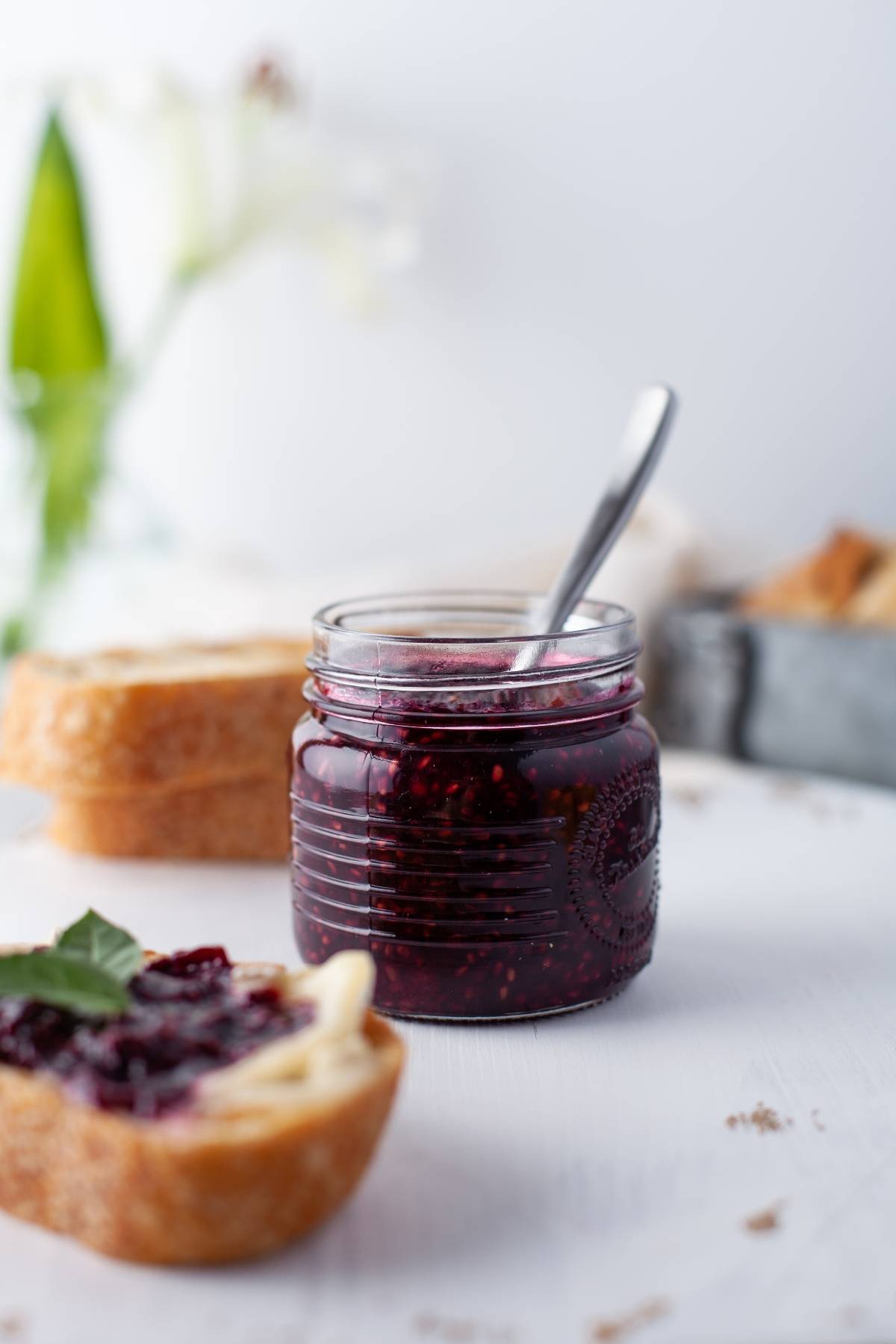 A jar of jam made with fresh berries and a spoon sits beside sliced bread, with a blurred flower in the background.