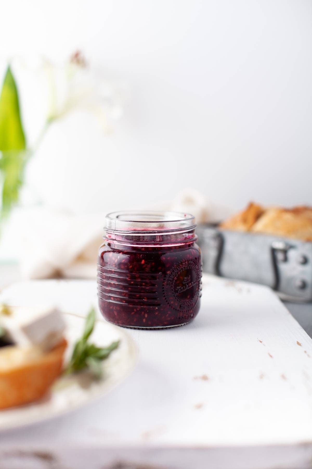 A jar of jam made with fresh berries sits on a white surface, with bread and a flower blurred in the background.