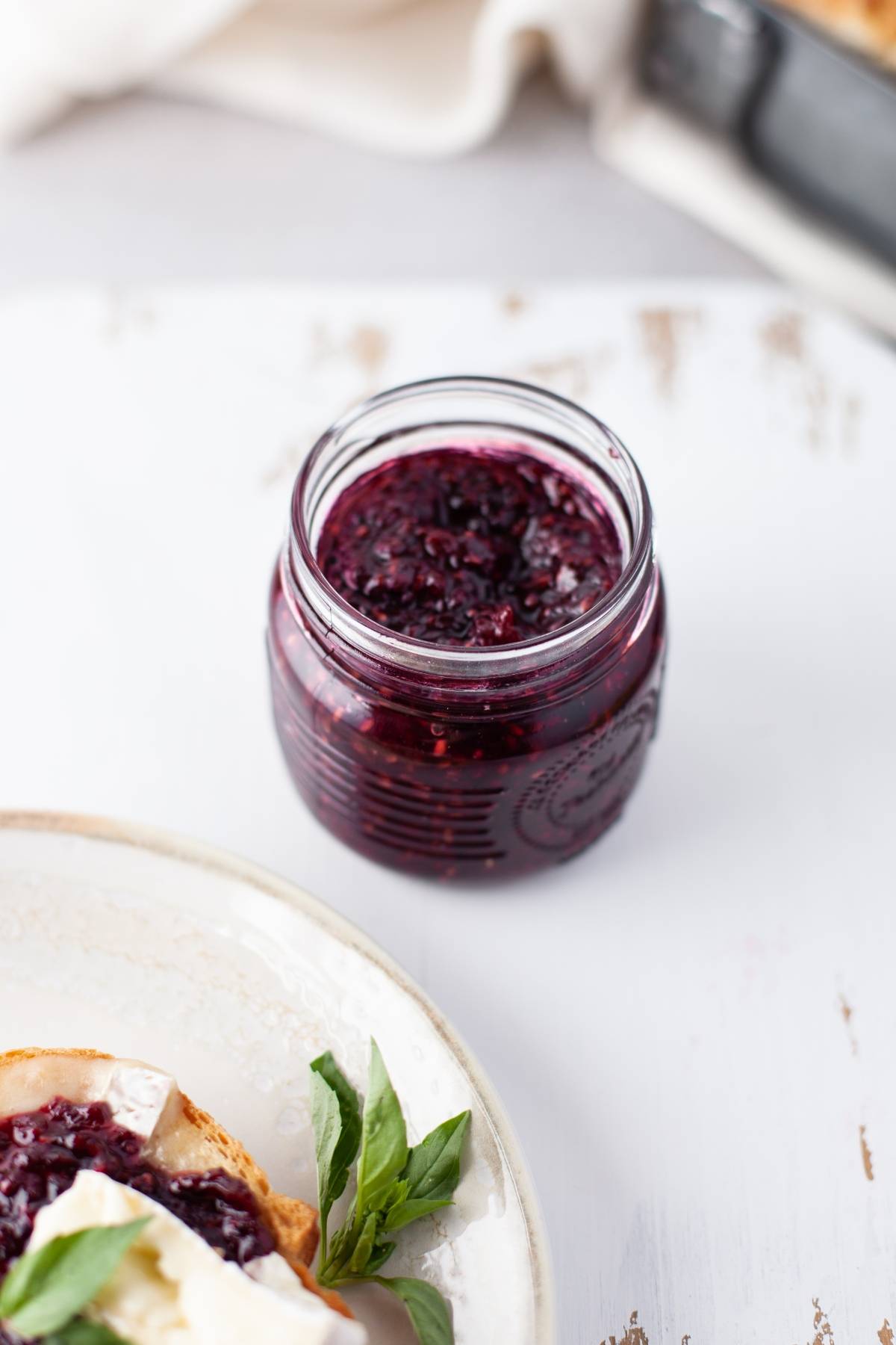 A small glass jar filled with dark berry jam made using the easiest jam recipe sits on a white surface next to a plate with bread.