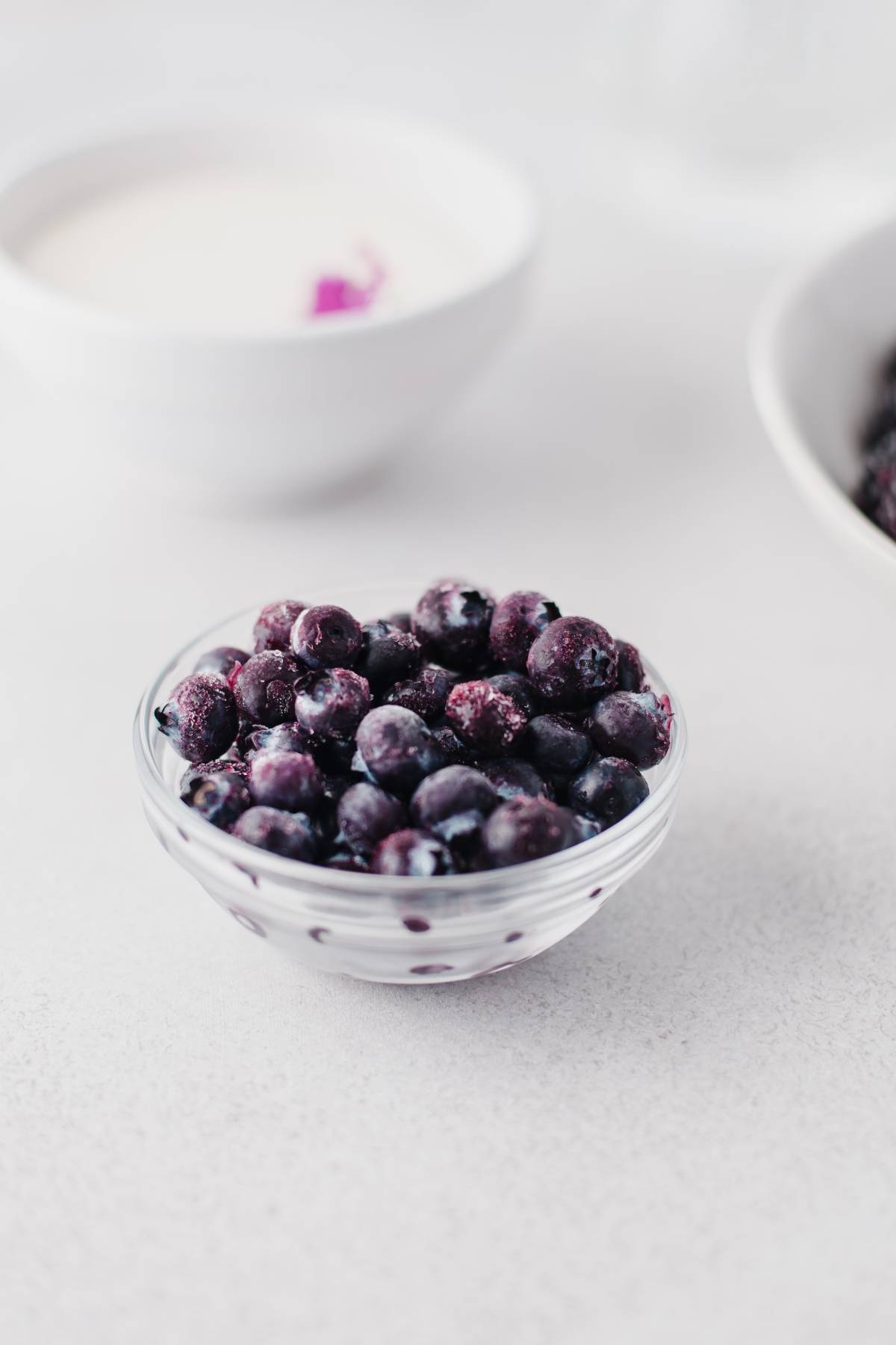 A glass bowl filled with frozen blueberries on a light surface, perfect for making sugar free blackberry jam, with bowls in the background.