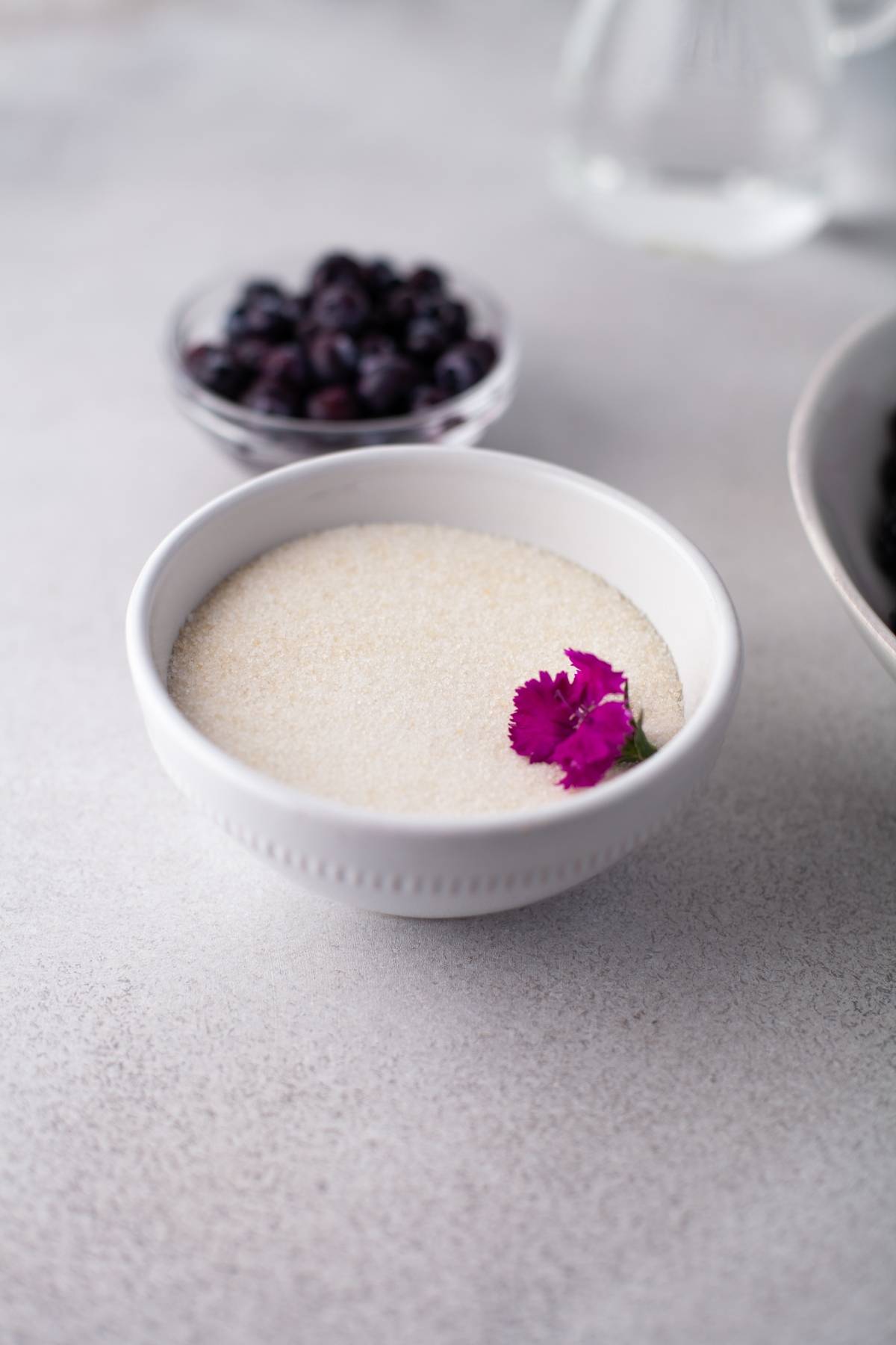 A white bowl of granulated sugar with a purple flower, with blueberries in a bowl in the background—perfect inspiration for the easiest jam recipe or a delicious sugar free blackberry jam made with fresh berries.