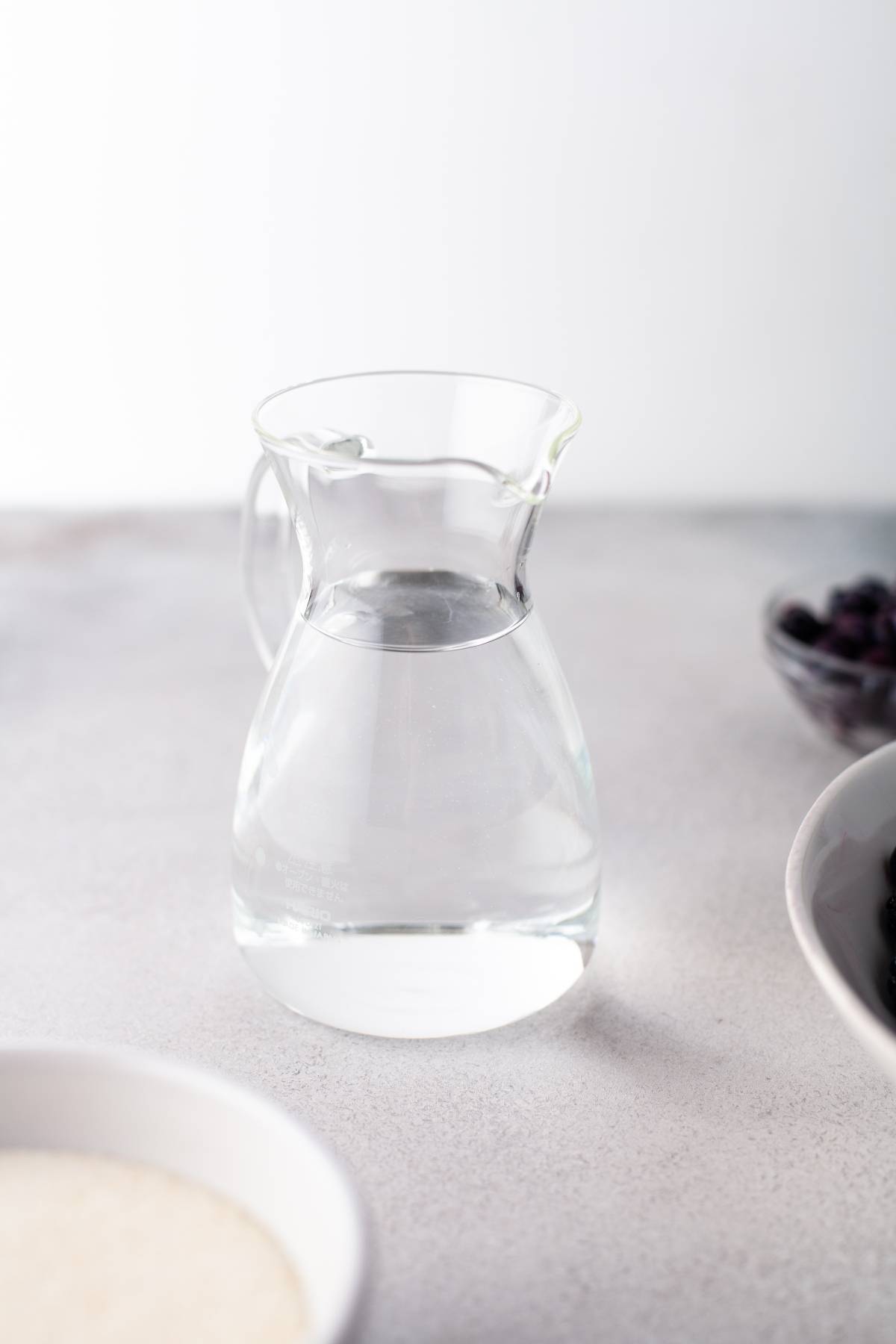A clear glass pitcher filled with water sits on a light gray surface, next to blurred bowls—perhaps ready for the easiest jam recipe or a sweet jam made with fresh berries.