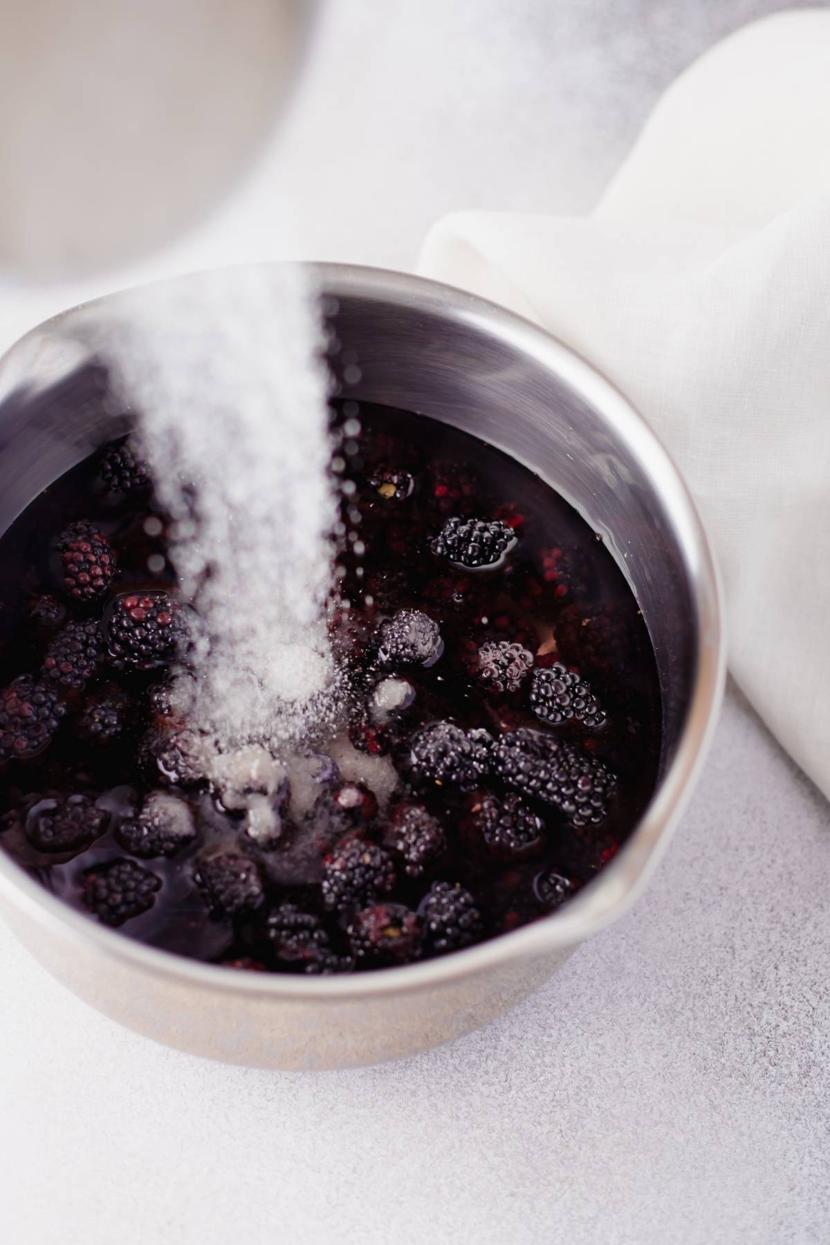 Sugar being poured into a metal bowl filled with blackberries and liquid, next to a white cloth—perfect for creating the easiest jam recipe made with fresh berries.