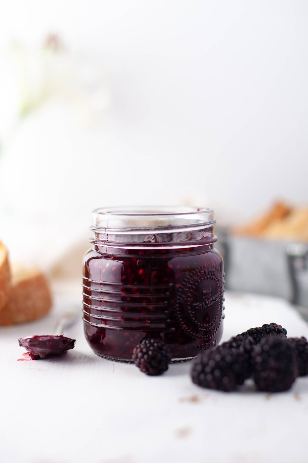 A jar of sugar free blackberry jam sits on a white surface with fresh blackberries and bread in the background, highlighting this healthy homemade jam recipe made with fresh berries.