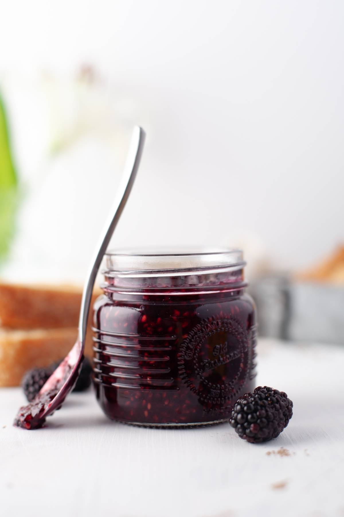 A jar of sugar free blackberry jam with a spoon resting on it, fresh blackberries, and bread in the background—perfect for a healthy homemade jam recipe.