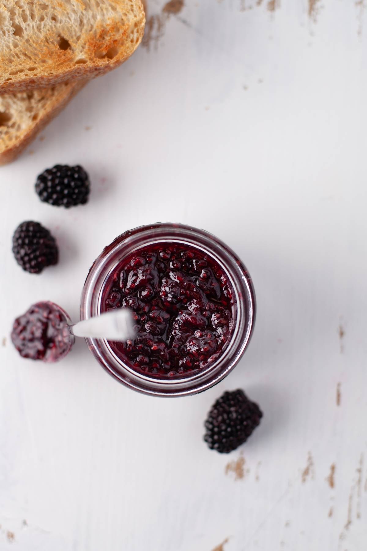 Jar of sugar free blackberry jam with a spoon, surrounded by blackberries and a slice of bread on a white surface—perfect for those seeking a healthy homemade jam recipe.