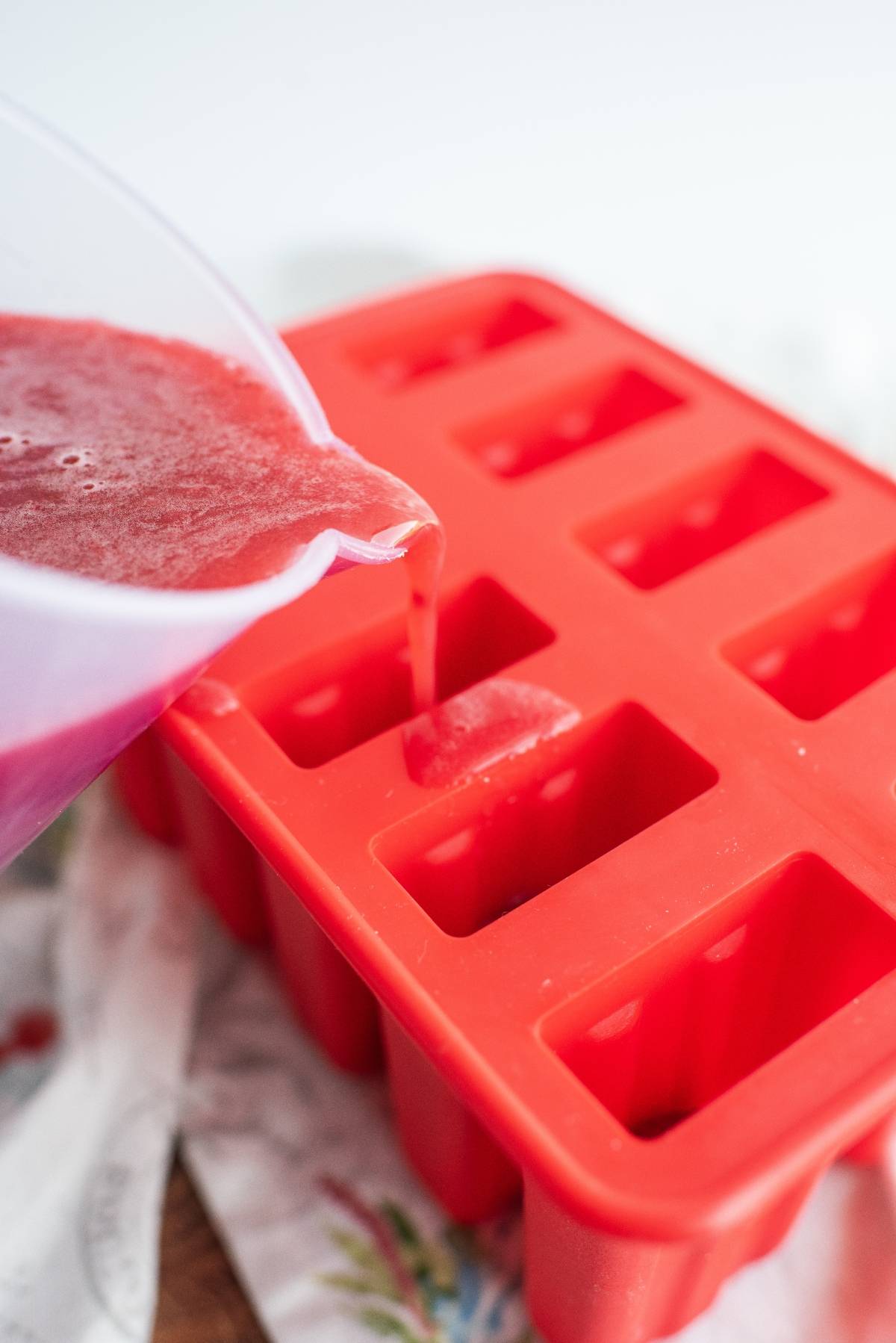 Red juice is being poured from a pitcher into a red silicone ice cube tray on a white surface, perfect for making Sweet Tea Raspberry Popsicles—refreshing sweet summer treats everyone will love.