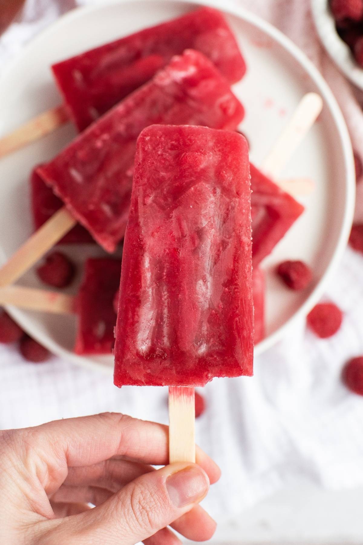 A hand holding a bright red Sweet Tea Raspberry Popsicle, with more sweet summer treats and fresh raspberries on a plate in the background.