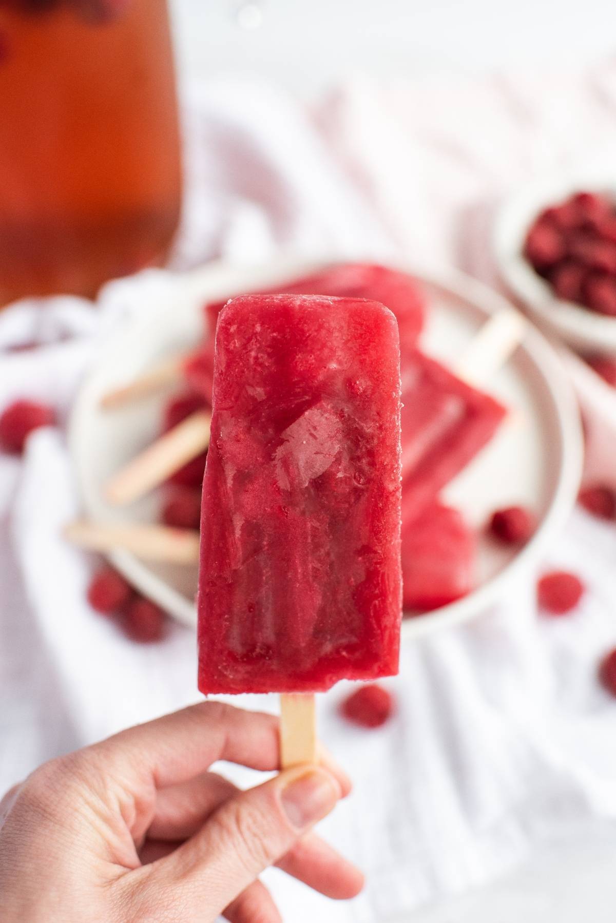 Hand holding a red popsicle with a plate of more popsicles and raspberries in the background—perfect sweet summer treats inspired by a raspberry ice pop recipe.