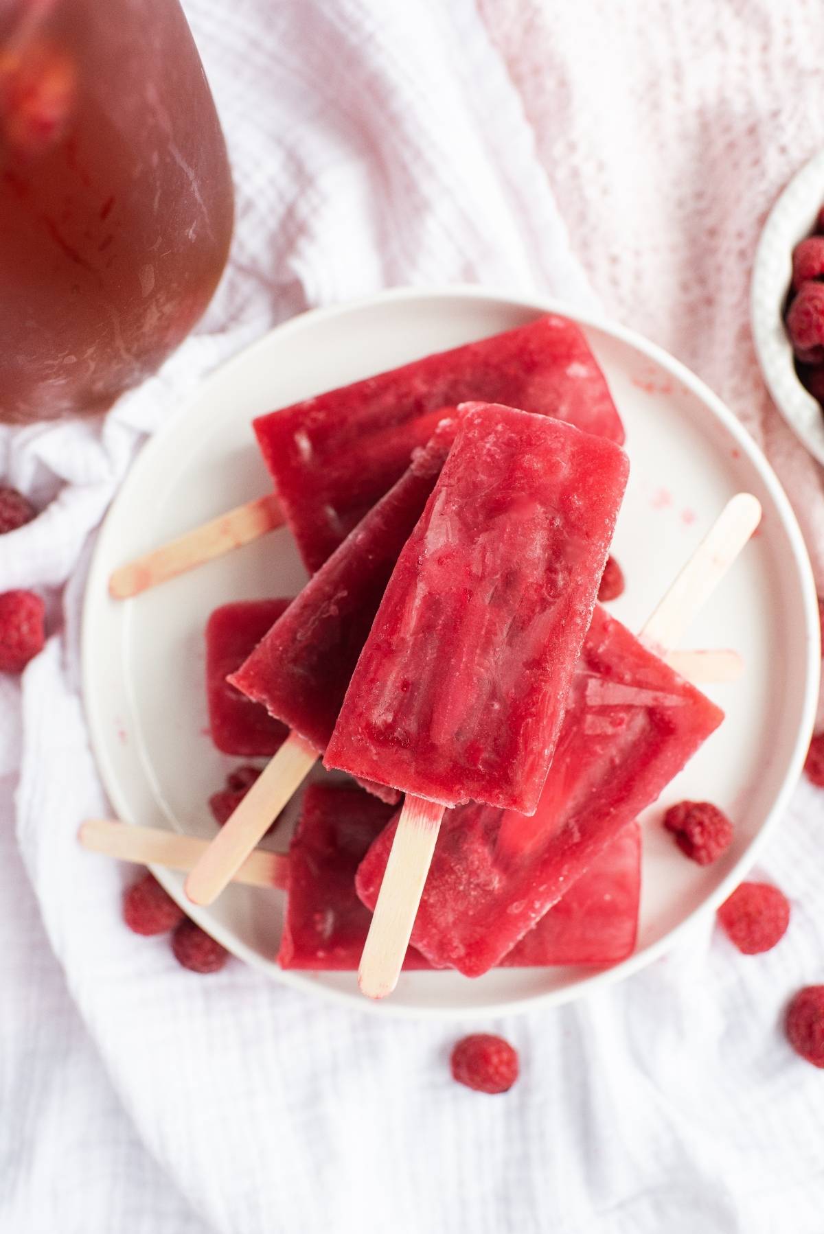 A plate of stacked Sweet Tea Raspberry Popsicles on a white cloth, with fresh raspberries scattered around—sweet summer treats made using frozen or fresh raspberries.